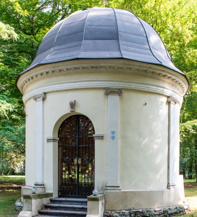 Zdjęcie nr 15: Kaplicę wzniesiono na rzucie koła, nakrywając jej wnętrze półkolistą kopułą. Ściany zewnętrzne zostały podzielone ośmioma pilastrami jońskimi, wspierającymi belkowanie. Powyżej belkowania biegnie profilowany gzyms koronujący, dekorowany fryzem kostkowym. Półokrągłe wejście usytuowano od zachodu. Prowadzi do niego pięć stopni. Wejście zamyka żelazna, klasycystyczna krata. Od wschodu w ścianie przepruto okulus, ujęty od wnętrza półokrągłym glifem w płytkiej, prostokątnej niszy zamkniętej półokrągło. Wnętrze pokryto ochrową polichromią, z wymalowanym białą kreską podziałem odzwierciedlającym artykulację zewnętrzną. Na ścianie umieszczono dwie czworoboczne płyty upamiętniające zmarłych pochowanych w krypcie. Wejście do krypty zamyka prostokątna, marmurowa płyta dekorowana aplikacją ze złoconego brązu, ukazującą trupią czaszkę ze skrzyżowanymi piszczelami umieszczoną nad antabą.