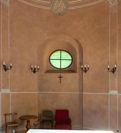 Zdjęcie nr 10: Kaplicę wzniesiono na rzucie koła, nakrywając jej wnętrze półkolistą kopułą. Ściany zewnętrzne zostały podzielone ośmioma pilastrami jońskimi, wspierającymi belkowanie. Powyżej belkowania biegnie profilowany gzyms koronujący, dekorowany fryzem kostkowym. Półokrągłe wejście usytuowano od zachodu. Prowadzi do niego pięć stopni. Wejście zamyka żelazna, klasycystyczna krata. Od wschodu w ścianie przepruto okulus, ujęty od wnętrza półokrągłym glifem w płytkiej, prostokątnej niszy zamkniętej półokrągło. Wnętrze pokryto ochrową polichromią, z wymalowanym białą kreską podziałem odzwierciedlającym artykulację zewnętrzną. Na ścianie umieszczono dwie czworoboczne płyty upamiętniające zmarłych pochowanych w krypcie. Wejście do krypty zamyka prostokątna, marmurowa płyta dekorowana aplikacją ze złoconego brązu, ukazującą trupią czaszkę ze skrzyżowanymi piszczelami umieszczoną nad antabą.