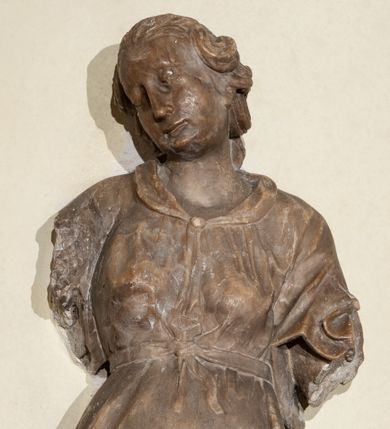 Zdjęcie nr 2: Przyścienna rzeźba ukazująca frontalnie młodą kobietę w całej postaci w kontrapoście umieszczona została na niskiej, czworobocznej podstawie. Postać ma lekko pochyloną głowę w lewo. Otaczają je falujące włosy spływające na plecy. Nie zachowały się ręce, które zostały ubite. Jej owalną twarz charakteryzuje wydatny nos oraz usta o pełnych wargach, a także spojrzenie skierowane w dół. Kobieta ubrana jest w zwiewną suknię przewiązaną w pasie wstążką. U dołu widać fragmenty bosych stóp. 