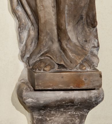 Zdjęcie nr 5: Przyścienna rzeźba ukazująca frontalnie młodą kobietę w całej postaci w kontrapoście umieszczona została na niskiej, czworobocznej podstawie. Postać ma lekko pochyloną głowę w lewo. Otaczają je falujące włosy spływające na plecy. Nie zachowały się ręce, które zostały ubite. Jej owalną twarz charakteryzuje wydatny nos oraz usta o pełnych wargach, a także spojrzenie skierowane w dół. Kobieta ubrana jest w zwiewną suknię przewiązaną w pasie wstążką. U dołu widać fragmenty bosych stóp. 