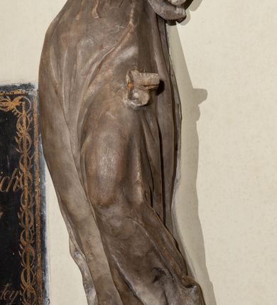 Zdjęcie nr 4: Przyścienna rzeźba ukazująca frontalnie młodą kobietę w całej postaci w kontrapoście umieszczona została na niskiej, czworobocznej podstawie. Postać ma lekko pochyloną głowę w lewo. Otaczają je falujące włosy spływające na plecy. Nie zachowały się ręce, które zostały ubite. Jej owalną twarz charakteryzuje wydatny nos oraz usta o pełnych wargach, a także spojrzenie skierowane w dół. Kobieta ubrana jest w zwiewną suknię przewiązaną w pasie wstążką. U dołu widać fragmenty bosych stóp. 