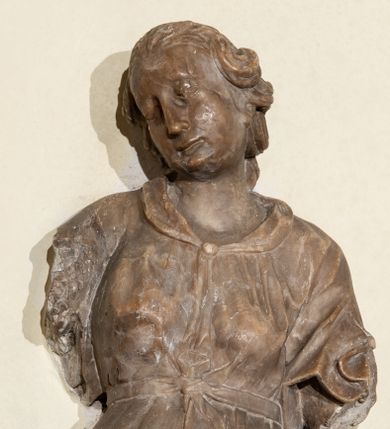 Zdjęcie nr 1: Przyścienna rzeźba ukazująca frontalnie młodą kobietę w całej postaci w kontrapoście umieszczona została na niskiej, czworobocznej podstawie. Postać ma lekko pochyloną głowę w lewo. Otaczają je falujące włosy spływające na plecy. Nie zachowały się ręce, które zostały ubite. Jej owalną twarz charakteryzuje wydatny nos oraz usta o pełnych wargach, a także spojrzenie skierowane w dół. Kobieta ubrana jest w zwiewną suknię przewiązaną w pasie wstążką. U dołu widać fragmenty bosych stóp. 