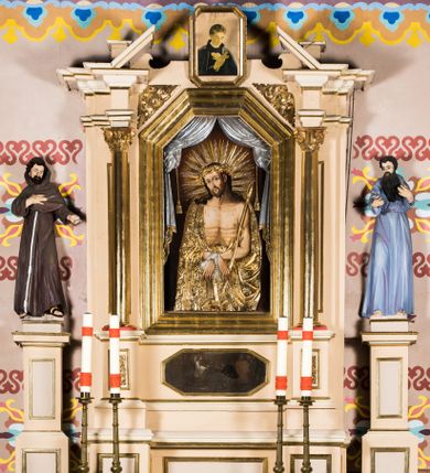 Zdjęcie nr 1: Przyścienny, jednoosiowy ołtarz uzyskał formę architektoniczną, jednokondygnacyjną. Stipes ołtarzowy otrzymał  kształt sarkofagowy z wybrzuszoną dolną częścią. Retabulum ustawiono na podwójnym cokole, ujmując kondygnacje parą kolumn o stylizowanych kapitelach kompozytowych. Podpory dźwigają odcinki belkowania z trójkątnym przerwanym przyczółkiem. W części środkowej usytuowano prostokątną wnękę zamkniętą od góry trójbocznie, obramienioną profilowaną, złoconą ramą. We wnęce zamontowano obraz Pana Jezusa Cierpiącego w typie Ecce Homo. Poniżej w drugiej strefie cokołu znalazł się obraz w kształcie leżącego prostokąta ze ściętymi narożami, przedstawiający cud z osłem św. Antoniego z Padwy. W partii przerwanego przyczółka zawieszono obraz w profilowanej ramie o ściętych narożach z przedstawieniem św. Stanisława Kostki. Nastawę ujmują trójstrefowe, smukłe cokoły z figurami św. Franciszka z Asyżu po lewej i świętego Pawła Pustelnika po prawej. Na cokołach i ponad ramą środkowego obrazu widać motyw ornamentu akantowego. 