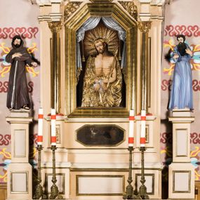 Zdjęcie nr 1: Przyścienny, jednoosiowy ołtarz uzyskał formę architektoniczną, jednokondygnacyjną. Stipes ołtarzowy otrzymał  kształt sarkofagowy z wybrzuszoną dolną częścią. Retabulum ustawiono na podwójnym cokole, ujmując kondygnacje parą kolumn o stylizowanych kapitelach kompozytowych. Podpory dźwigają odcinki belkowania z trójkątnym przerwanym przyczółkiem. W części środkowej usytuowano prostokątną wnękę zamkniętą od góry trójbocznie, obramienioną profilowaną, złoconą ramą. We wnęce zamontowano obraz Pana Jezusa Cierpiącego w typie Ecce Homo. Poniżej w drugiej strefie cokołu znalazł się obraz w kształcie leżącego prostokąta ze ściętymi narożami, przedstawiający cud z osłem św. Antoniego z Padwy. W partii przerwanego przyczółka zawieszono obraz w profilowanej ramie o ściętych narożach z przedstawieniem św. Stanisława Kostki. Nastawę ujmują trójstrefowe, smukłe cokoły z figurami św. Franciszka z Asyżu po lewej i świętego Pawła Pustelnika po prawej. Na cokołach i ponad ramą środkowego obrazu widać motyw ornamentu akantowego. 