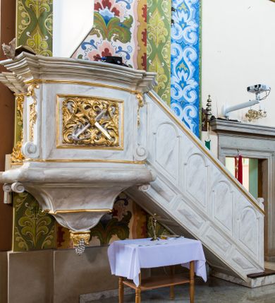 Zdjęcie nr 1: Ambona przyścienna, wisząca, złożona z kosza i schodów z balustradą. Kosz jest  czworoboczny, o kształcie wklęsło-wypukłym i ściętych narożach dekorowanych spływami zakończonymi ślimacznicami; na nich różnorodny ornament rocaille’owy. Ścianki kosza wypełnione płycinami, boczna z ornamentem rocaille’owym, frontowa z kompozycją złożoną ze skrzyżowanych krzyża i kotwicy, na przecięciu których widnieje gorejące serce w otoczeniu wici akantowej i gałązek róż. Kosz wieńczy mocno wysunięty i bogato profilowany gzyms stanowiący parapet. Od dołu kończy go wałek z uskokiem na narożu i podwieszenie w formie wklęsło-wypukłego odwróconego stożka z uskokiem pośrodku, zakończonego kiścią winogron. Z prawej strony do kosza dochodzą schody z balustradą zdobioną płycinami: w dolnej części prostokątnymi, wypełnionymi rautami, a w górnej w kształcie stojących prostokątów o skośnych dolnych krawędziach i zamkniętych łukami nadwieszonymi. Struktura marmoryzowana na kolor biało-szary, profile i ornamenty złocone oraz srebrzone. 