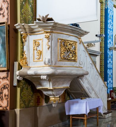 Zdjęcie nr 2: Ambona przyścienna, wisząca, złożona z kosza i schodów z balustradą. Kosz jest  czworoboczny, o kształcie wklęsło-wypukłym i ściętych narożach dekorowanych spływami zakończonymi ślimacznicami; na nich różnorodny ornament rocaille’owy. Ścianki kosza wypełnione płycinami, boczna z ornamentem rocaille’owym, frontowa z kompozycją złożoną ze skrzyżowanych krzyża i kotwicy, na przecięciu których widnieje gorejące serce w otoczeniu wici akantowej i gałązek róż. Kosz wieńczy mocno wysunięty i bogato profilowany gzyms stanowiący parapet. Od dołu kończy go wałek z uskokiem na narożu i podwieszenie w formie wklęsło-wypukłego odwróconego stożka z uskokiem pośrodku, zakończonego kiścią winogron. Z prawej strony do kosza dochodzą schody z balustradą zdobioną płycinami: w dolnej części prostokątnymi, wypełnionymi rautami, a w górnej w kształcie stojących prostokątów o skośnych dolnych krawędziach i zamkniętych łukami nadwieszonymi. Struktura marmoryzowana na kolor biało-szary, profile i ornamenty złocone oraz srebrzone. 