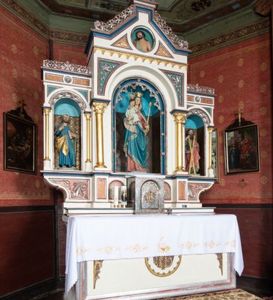 Zdjęcie nr 7: Ołtarz architektoniczny, przyścienny, jednokondygnacyjny, trójosiowy, ze zwieńczeniem. Ołtarz prostopadłościenny z antepedium zdobionym polichromowanym motywem liści akantu po bokach, pośrodku medalion z monogramem chrystologicznym. Tabernakulum w formie prostopadłościennej szafki z drzwiczkami zamkniętymi łukiem półkolistym ujętym fryzem arkadkowym, zdobionych monogramem chrystologicznym. Retabulum na niewysokim, zryzalitowanym cokole o zakończeniach w formie konsoli. Osi głównej części nastawy są w formie półkolistych arkad z niszami, wspartych parami kolumn o kapitelach kompozytowych, przy czym środkowa jest większa względem bocznych dźwigających belkowanie. Zwieńczenie części środkowej retabulum przybiera formę zbliżoną do trójkątnego przyczółka. Pola cokołu retabulum zdobi polichromowana i płaskorzeźbiona ornamentyka roślinna. Ścianki niszy pokryte są dekoracją malarską naśladującą wzorzyste tkaniny, a w górnej partii błękitne, gwieździste niebo. Łuki nisz ujmuje ażurowy fryz arkadkowy, dodatkowo obramienie niszy głównej z motywem rozetek. W przyłuczach dekoracja w formie wici roślinnej. Krawędź attyki oraz zwieńczenia zdobi ażurowa wić roślinna. W polu części środkowej znajduje się figura Madonny z Dzieciątkiem, w arkadzie po prawej stronie ołtarza św. Andrzeja, po lewej św. Piotra. Struktura ołtarza utrzymana jest w kolorach białym, niebieskim, zielonym i jasnoczerwonym, detal architektoniczny i ornamentyka są złocone.        