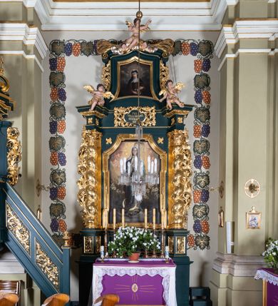 Zdjęcie nr 2: Nastawa architektoniczna, jednoosiowa, jednokondygnacyjna ze zwieńczeniem, ustawiona na dwustrefowym cokole z wyodrębnionymi dwoma prostopadłościanami. Ołtarz prostopadłościenny, z drewnianą mensą. Antepedium ujęte profilowaną, złoconą ramą, na tle fioletowego pola mandorla z chrystogramem Chi Rho oraz literami alfa i omega, w glorii promienistej. Cokół w górnej strefie dekorowany płycinami wypełnionymi akantem. Nastawa flankowana parą pilastrów o kanelowanych trzonach i stojących przed nimi kolumn o kapitelach kompozytowych i kręconych trzonach, oplecionych akantem, ustawionych pod kątem na zewnątrz, dźwigających pełne belkowanie, wyłamane na ich osi, o fryzie dekorowanym rozetami, z siedzącymi na nim puttami. Pole centralne niemal w całości wypełnione wnęką ujętą profilowaną ramą w formie stojącego prostokąta o wklęsłych górnych narożach. We wnęce obraz z przedstawieniem Niepokalanego Poczęcia Matki Boskiej, na zasuwie św. Anna Samotrzeć. Nad obrazem, w polu belkowania kartusz utworzony z akantu, girland i zwisów z napisem „POD / TWOJĄ OBRONĘ / UCIEKAMY / SIĘ”. Kondygnacja ujęta uszami utworzonymi z akantu. Zwieńczenie w formie stojącego prostokąta o lekko wklęsłych bokach, zamknięte fragmentem segmentowego gzymsu zakończonego wolutami, dekorowanego obłokami, liśćmi oraz figurą putta i uskrzydlonych główek anielskich. W polu obraz św. Róży z Viterbo w formie zbliżonej do trapezu o wklęsłych podstawach, ujęty profilowaną ramą. Zwieńczenie flankowane uszami z akantu i kampanuli. Struktura polichromowana w kolorze zielonym, ze złoconymi kolumnami, profilami i ornamentyką. 