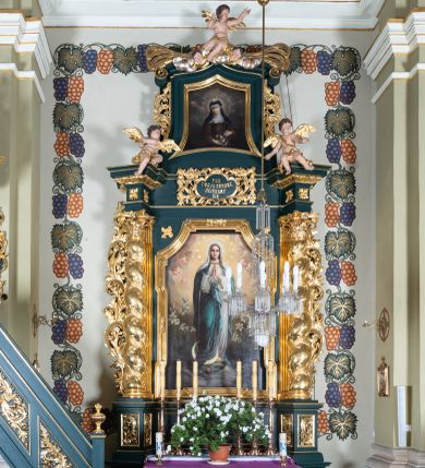 Zdjęcie nr 1: Nastawa architektoniczna, jednoosiowa, jednokondygnacyjna ze zwieńczeniem, ustawiona na dwustrefowym cokole z wyodrębnionymi dwoma prostopadłościanami. Ołtarz prostopadłościenny, z drewnianą mensą. Antepedium ujęte profilowaną, złoconą ramą, na tle fioletowego pola mandorla z chrystogramem Chi Rho oraz literami alfa i omega, w glorii promienistej. Cokół w górnej strefie dekorowany płycinami wypełnionymi akantem. Nastawa flankowana parą pilastrów o kanelowanych trzonach i stojących przed nimi kolumn o kapitelach kompozytowych i kręconych trzonach, oplecionych akantem, ustawionych pod kątem na zewnątrz, dźwigających pełne belkowanie, wyłamane na ich osi, o fryzie dekorowanym rozetami, z siedzącymi na nim puttami. Pole centralne niemal w całości wypełnione wnęką ujętą profilowaną ramą w formie stojącego prostokąta o wklęsłych górnych narożach. We wnęce obraz z przedstawieniem Niepokalanego Poczęcia Matki Boskiej, na zasuwie św. Anna Samotrzeć. Nad obrazem, w polu belkowania kartusz utworzony z akantu, girland i zwisów z napisem „POD / TWOJĄ OBRONĘ / UCIEKAMY / SIĘ”. Kondygnacja ujęta uszami utworzonymi z akantu. Zwieńczenie w formie stojącego prostokąta o lekko wklęsłych bokach, zamknięte fragmentem segmentowego gzymsu zakończonego wolutami, dekorowanego obłokami, liśćmi oraz figurą putta i uskrzydlonych główek anielskich. W polu obraz św. Róży z Viterbo w formie zbliżonej do trapezu o wklęsłych podstawach, ujęty profilowaną ramą. Zwieńczenie flankowane uszami z akantu i kampanuli. Struktura polichromowana w kolorze zielonym, ze złoconymi kolumnami, profilami i ornamentyką. 