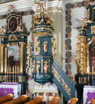 Zdjęcie nr 2: Ambona przyścienna, wisząca, z dostępem w formie jednobiegowych schodów. Kosz na planie ściętego ośmioboku, z podwieszeniem w formie spłaszczonego, ośmiobocznego wałka, zakończonego szyszką. Kosz na wysokim cokole, ścianki wydzielone pilastrami dekorowanymi muszlami i zwisami kwiatowymi, dźwigającymi pełne belkowanie z silnie wysuniętym gzymsem tworzącym parapet, dekorowanym ornamentem roślinnym. W polu ścianek nisze z konchami i figurami czterech ewangelistów, stojącymi na wspornikach. Balustrada schodów pełna, dekorowana płycinami wypełnionymi akantem, zakończona filarem z wazonem. Zaplecek zakończony gzymsem, ujęty uszami utworzonymi z suchego akantu. W polu płycina w formie stojącego prostokąta o wklęsłych narożach, w niej nisza z konchą, na tle której na wsporniku ustawiona figura Chrystusa. Baldachim na planie ściętego ośmioboku, w formie belkowania, wyłamanego na narożach. Na nim, na niskiej podstawie osiem kabłąków dekorowanych cokolikami z wazonami, podtrzymujących spłaszczoną kulę, na której stoi figura anioła z banderolą z napisem „KTO WAS SŁUCHA MNIE SŁUCHA”. Na podniebieniu baldachimu rzeźba gołębicy Ducha Świętego w glorii promienistej. Struktura polichromowana w kolorze zielonym, ze złoconymi profilami i ornamentyką. 

Figura anioła ukazana frontalnie, w kontrapoście, z głową skierowaną w lewo, obiema rękami trzyma banderolę. Twarz ma pociągłą, o migdałowatych oczach, dużym nosie i rozchylonych ustach, okoloną krótkimi, kręconymi, rozwianymi włosami. Anioł ubrany jest w długą, silnie drapowaną, złoconą tunikę, przepasaną w talii, odsłaniającą prawe ramię oraz obie nogi. Skrzydła ma złożone po bokach postaci. 

Figura Chrystusa ukazana frontalnie, w kontrapoście, prawą rękę unosi w geście błogosławieństwa, w lewej trzyma jabłko królewskie. Twarz ma szczupłą, o migdałowatych oczach, dużym nosie i wąskich ustach, okoloną brązową brodą i włosami sięgającymi ramion. Chrystus ubrany jest w przylegającą do ciała, złoconą tunikę, przepasaną w talii oraz złocony płaszcz o czerwonej podszewce, przerzucony diagonalnie przez lewe ramię. 

Figura św. Jana ukazana frontalnie, w kontrapoście, przechylona w lewo, prawą rękę wyciąga do przodu, lewą podtrzymuje otwartą księgę. Twarz ma owalną, o migdałowatych oczach, dużym nosie i wąskich ustach, okoloną brązowymi włosami sięgającymi ramion. Święty ubrany jest w przylegającą do ciała, złoconą tunikę, przepasaną w talii oraz złocony płaszcz o czerwonej podszewce, przerzucony diagonalnie przez lewe ramię, zawieszony na taśmie z przodu postaci. 

Figura św. Łukasza ukazana frontalnie, w kontrapoście, przechylona w lewo, prawą rękę unosi na tle piersi, lewą podtrzymuje otwartą księgę. Twarz ma owalną, o migdałowatych oczach, dużym nosie i wąskich ustach, okoloną brązową długą brodą i krótkimi włosami. Święty ubrany jest w przylegającą do ciała, złoconą tunikę, przepasaną w talii oraz złocony płaszcz o zielonej podszewce, przerzucony diagonalnie przez lewe ramię. Przy prawej nodze świętego głowa wołu. 

Figura św. Marka ukazana frontalnie, w kontrapoście, przechylona w lewo, z głową zwróconą w lewo, prawą rękę wyciąga w bok, lewą przytrzymuje przy boku księgę. Twarz ma owalną, o migdałowatych oczach, dużym nosie i wąskich ustach, okoloną gęstą, brązową brodą i krótkimi, kręconymi włosami. Święty ubrany jest w przylegającą do ciała, złoconą tunikę, przepasaną w talii oraz złocony płaszcz o jasnozielonej podszewce, zarzucony na ramiona i zawiązany z przodu. Przy lewej nodze postaci głowa lwa. 

Figura św. Mateusza ukazana frontalnie, w kontrapoście, lewą rękę wyciąga w bok, prawą przytrzymuje przy boku księgę. Twarz ma owalną, o migdałowatych oczach, dużym nosie i wąskich ustach, okoloną długą, brązową brodą i krótkimi włosami. Święty ubrany jest w przylegającą do ciała, złoconą tunikę, przepasaną w talii szarfą oraz złocony płaszcz o zielonej podszewce, zarzucony na ramiona i opadający po bokach. Przed świętym figurka aniołka trzymającego miskę. 

