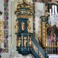 Zdjęcie nr 1: Ambona przyścienna, wisząca, z dostępem w formie jednobiegowych schodów. Kosz na planie ściętego ośmioboku, z podwieszeniem w formie spłaszczonego, ośmiobocznego wałka, zakończonego szyszką. Kosz na wysokim cokole, ścianki wydzielone pilastrami dekorowanymi muszlami i zwisami kwiatowymi, dźwigającymi pełne belkowanie z silnie wysuniętym gzymsem tworzącym parapet, dekorowanym ornamentem roślinnym. W polu ścianek nisze z konchami i figurami czterech ewangelistów, stojącymi na wspornikach. Balustrada schodów pełna, dekorowana płycinami wypełnionymi akantem, zakończona filarem z wazonem. Zaplecek zakończony gzymsem, ujęty uszami utworzonymi z suchego akantu. W polu płycina w formie stojącego prostokąta o wklęsłych narożach, w niej nisza z konchą, na tle której na wsporniku ustawiona figura Chrystusa. Baldachim na planie ściętego ośmioboku, w formie belkowania, wyłamanego na narożach. Na nim, na niskiej podstawie osiem kabłąków dekorowanych cokolikami z wazonami, podtrzymujących spłaszczoną kulę, na której stoi figura anioła z banderolą z napisem „KTO WAS SŁUCHA MNIE SŁUCHA”. Na podniebieniu baldachimu rzeźba gołębicy Ducha Świętego w glorii promienistej. Struktura polichromowana w kolorze zielonym, ze złoconymi profilami i ornamentyką. 

Figura anioła ukazana frontalnie, w kontrapoście, z głową skierowaną w lewo, obiema rękami trzyma banderolę. Twarz ma pociągłą, o migdałowatych oczach, dużym nosie i rozchylonych ustach, okoloną krótkimi, kręconymi, rozwianymi włosami. Anioł ubrany jest w długą, silnie drapowaną, złoconą tunikę, przepasaną w talii, odsłaniającą prawe ramię oraz obie nogi. Skrzydła ma złożone po bokach postaci. 

Figura Chrystusa ukazana frontalnie, w kontrapoście, prawą rękę unosi w geście błogosławieństwa, w lewej trzyma jabłko królewskie. Twarz ma szczupłą, o migdałowatych oczach, dużym nosie i wąskich ustach, okoloną brązową brodą i włosami sięgającymi ramion. Chrystus ubrany jest w przylegającą do ciała, złoconą tunikę, przepasaną w talii oraz złocony płaszcz o czerwonej podszewce, przerzucony diagonalnie przez lewe ramię. 

Figura św. Jana ukazana frontalnie, w kontrapoście, przechylona w lewo, prawą rękę wyciąga do przodu, lewą podtrzymuje otwartą księgę. Twarz ma owalną, o migdałowatych oczach, dużym nosie i wąskich ustach, okoloną brązowymi włosami sięgającymi ramion. Święty ubrany jest w przylegającą do ciała, złoconą tunikę, przepasaną w talii oraz złocony płaszcz o czerwonej podszewce, przerzucony diagonalnie przez lewe ramię, zawieszony na taśmie z przodu postaci. 

Figura św. Łukasza ukazana frontalnie, w kontrapoście, przechylona w lewo, prawą rękę unosi na tle piersi, lewą podtrzymuje otwartą księgę. Twarz ma owalną, o migdałowatych oczach, dużym nosie i wąskich ustach, okoloną brązową długą brodą i krótkimi włosami. Święty ubrany jest w przylegającą do ciała, złoconą tunikę, przepasaną w talii oraz złocony płaszcz o zielonej podszewce, przerzucony diagonalnie przez lewe ramię. Przy prawej nodze świętego głowa wołu. 

Figura św. Marka ukazana frontalnie, w kontrapoście, przechylona w lewo, z głową zwróconą w lewo, prawą rękę wyciąga w bok, lewą przytrzymuje przy boku księgę. Twarz ma owalną, o migdałowatych oczach, dużym nosie i wąskich ustach, okoloną gęstą, brązową brodą i krótkimi, kręconymi włosami. Święty ubrany jest w przylegającą do ciała, złoconą tunikę, przepasaną w talii oraz złocony płaszcz o jasnozielonej podszewce, zarzucony na ramiona i zawiązany z przodu. Przy lewej nodze postaci głowa lwa. 

Figura św. Mateusza ukazana frontalnie, w kontrapoście, lewą rękę wyciąga w bok, prawą przytrzymuje przy boku księgę. Twarz ma owalną, o migdałowatych oczach, dużym nosie i wąskich ustach, okoloną długą, brązową brodą i krótkimi włosami. Święty ubrany jest w przylegającą do ciała, złoconą tunikę, przepasaną w talii szarfą oraz złocony płaszcz o zielonej podszewce, zarzucony na ramiona i opadający po bokach. Przed świętym figurka aniołka trzymającego miskę. 
