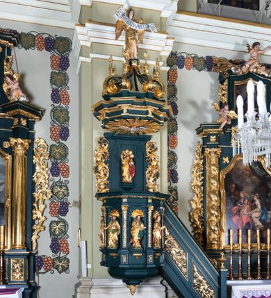 Zdjęcie nr 1: Ambona przyścienna, wisząca, z dostępem w formie jednobiegowych schodów. Kosz na planie ściętego ośmioboku, z podwieszeniem w formie spłaszczonego, ośmiobocznego wałka, zakończonego szyszką. Kosz na wysokim cokole, ścianki wydzielone pilastrami dekorowanymi muszlami i zwisami kwiatowymi, dźwigającymi pełne belkowanie z silnie wysuniętym gzymsem tworzącym parapet, dekorowanym ornamentem roślinnym. W polu ścianek nisze z konchami i figurami czterech ewangelistów, stojącymi na wspornikach. Balustrada schodów pełna, dekorowana płycinami wypełnionymi akantem, zakończona filarem z wazonem. Zaplecek zakończony gzymsem, ujęty uszami utworzonymi z suchego akantu. W polu płycina w formie stojącego prostokąta o wklęsłych narożach, w niej nisza z konchą, na tle której na wsporniku ustawiona figura Chrystusa. Baldachim na planie ściętego ośmioboku, w formie belkowania, wyłamanego na narożach. Na nim, na niskiej podstawie osiem kabłąków dekorowanych cokolikami z wazonami, podtrzymujących spłaszczoną kulę, na której stoi figura anioła z banderolą z napisem „KTO WAS SŁUCHA MNIE SŁUCHA”. Na podniebieniu baldachimu rzeźba gołębicy Ducha Świętego w glorii promienistej. Struktura polichromowana w kolorze zielonym, ze złoconymi profilami i ornamentyką. 

Figura anioła ukazana frontalnie, w kontrapoście, z głową skierowaną w lewo, obiema rękami trzyma banderolę. Twarz ma pociągłą, o migdałowatych oczach, dużym nosie i rozchylonych ustach, okoloną krótkimi, kręconymi, rozwianymi włosami. Anioł ubrany jest w długą, silnie drapowaną, złoconą tunikę, przepasaną w talii, odsłaniającą prawe ramię oraz obie nogi. Skrzydła ma złożone po bokach postaci. 

Figura Chrystusa ukazana frontalnie, w kontrapoście, prawą rękę unosi w geście błogosławieństwa, w lewej trzyma jabłko królewskie. Twarz ma szczupłą, o migdałowatych oczach, dużym nosie i wąskich ustach, okoloną brązową brodą i włosami sięgającymi ramion. Chrystus ubrany jest w przylegającą do ciała, złoconą tunikę, przepasaną w talii oraz złocony płaszcz o czerwonej podszewce, przerzucony diagonalnie przez lewe ramię. 

Figura św. Jana ukazana frontalnie, w kontrapoście, przechylona w lewo, prawą rękę wyciąga do przodu, lewą podtrzymuje otwartą księgę. Twarz ma owalną, o migdałowatych oczach, dużym nosie i wąskich ustach, okoloną brązowymi włosami sięgającymi ramion. Święty ubrany jest w przylegającą do ciała, złoconą tunikę, przepasaną w talii oraz złocony płaszcz o czerwonej podszewce, przerzucony diagonalnie przez lewe ramię, zawieszony na taśmie z przodu postaci. 

Figura św. Łukasza ukazana frontalnie, w kontrapoście, przechylona w lewo, prawą rękę unosi na tle piersi, lewą podtrzymuje otwartą księgę. Twarz ma owalną, o migdałowatych oczach, dużym nosie i wąskich ustach, okoloną brązową długą brodą i krótkimi włosami. Święty ubrany jest w przylegającą do ciała, złoconą tunikę, przepasaną w talii oraz złocony płaszcz o zielonej podszewce, przerzucony diagonalnie przez lewe ramię. Przy prawej nodze świętego głowa wołu. 

Figura św. Marka ukazana frontalnie, w kontrapoście, przechylona w lewo, z głową zwróconą w lewo, prawą rękę wyciąga w bok, lewą przytrzymuje przy boku księgę. Twarz ma owalną, o migdałowatych oczach, dużym nosie i wąskich ustach, okoloną gęstą, brązową brodą i krótkimi, kręconymi włosami. Święty ubrany jest w przylegającą do ciała, złoconą tunikę, przepasaną w talii oraz złocony płaszcz o jasnozielonej podszewce, zarzucony na ramiona i zawiązany z przodu. Przy lewej nodze postaci głowa lwa. 

Figura św. Mateusza ukazana frontalnie, w kontrapoście, lewą rękę wyciąga w bok, prawą przytrzymuje przy boku księgę. Twarz ma owalną, o migdałowatych oczach, dużym nosie i wąskich ustach, okoloną długą, brązową brodą i krótkimi włosami. Święty ubrany jest w przylegającą do ciała, złoconą tunikę, przepasaną w talii szarfą oraz złocony płaszcz o zielonej podszewce, zarzucony na ramiona i opadający po bokach. Przed świętym figurka aniołka trzymającego miskę. 
