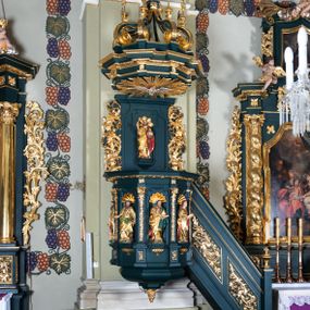 Zdjęcie nr 1: Ambona przyścienna, wisząca, z dostępem w formie jednobiegowych schodów. Kosz na planie ściętego ośmioboku, z podwieszeniem w formie spłaszczonego, ośmiobocznego wałka, zakończonego szyszką. Kosz na wysokim cokole, ścianki wydzielone pilastrami dekorowanymi muszlami i zwisami kwiatowymi, dźwigającymi pełne belkowanie z silnie wysuniętym gzymsem tworzącym parapet, dekorowanym ornamentem roślinnym. W polu ścianek nisze z konchami i figurami czterech ewangelistów, stojącymi na wspornikach. Balustrada schodów pełna, dekorowana płycinami wypełnionymi akantem, zakończona filarem z wazonem. Zaplecek zakończony gzymsem, ujęty uszami utworzonymi z suchego akantu. W polu płycina w formie stojącego prostokąta o wklęsłych narożach, w niej nisza z konchą, na tle której na wsporniku ustawiona figura Chrystusa. Baldachim na planie ściętego ośmioboku, w formie belkowania, wyłamanego na narożach. Na nim, na niskiej podstawie osiem kabłąków dekorowanych cokolikami z wazonami, podtrzymujących spłaszczoną kulę, na której stoi figura anioła z banderolą z napisem „KTO WAS SŁUCHA MNIE SŁUCHA”. Na podniebieniu baldachimu rzeźba gołębicy Ducha Świętego w glorii promienistej. Struktura polichromowana w kolorze zielonym, ze złoconymi profilami i ornamentyką. 

Figura anioła ukazana frontalnie, w kontrapoście, z głową skierowaną w lewo, obiema rękami trzyma banderolę. Twarz ma pociągłą, o migdałowatych oczach, dużym nosie i rozchylonych ustach, okoloną krótkimi, kręconymi, rozwianymi włosami. Anioł ubrany jest w długą, silnie drapowaną, złoconą tunikę, przepasaną w talii, odsłaniającą prawe ramię oraz obie nogi. Skrzydła ma złożone po bokach postaci. 

Figura Chrystusa ukazana frontalnie, w kontrapoście, prawą rękę unosi w geście błogosławieństwa, w lewej trzyma jabłko królewskie. Twarz ma szczupłą, o migdałowatych oczach, dużym nosie i wąskich ustach, okoloną brązową brodą i włosami sięgającymi ramion. Chrystus ubrany jest w przylegającą do ciała, złoconą tunikę, przepasaną w talii oraz złocony płaszcz o czerwonej podszewce, przerzucony diagonalnie przez lewe ramię. 

Figura św. Jana ukazana frontalnie, w kontrapoście, przechylona w lewo, prawą rękę wyciąga do przodu, lewą podtrzymuje otwartą księgę. Twarz ma owalną, o migdałowatych oczach, dużym nosie i wąskich ustach, okoloną brązowymi włosami sięgającymi ramion. Święty ubrany jest w przylegającą do ciała, złoconą tunikę, przepasaną w talii oraz złocony płaszcz o czerwonej podszewce, przerzucony diagonalnie przez lewe ramię, zawieszony na taśmie z przodu postaci. 

Figura św. Łukasza ukazana frontalnie, w kontrapoście, przechylona w lewo, prawą rękę unosi na tle piersi, lewą podtrzymuje otwartą księgę. Twarz ma owalną, o migdałowatych oczach, dużym nosie i wąskich ustach, okoloną brązową długą brodą i krótkimi włosami. Święty ubrany jest w przylegającą do ciała, złoconą tunikę, przepasaną w talii oraz złocony płaszcz o zielonej podszewce, przerzucony diagonalnie przez lewe ramię. Przy prawej nodze świętego głowa wołu. 

Figura św. Marka ukazana frontalnie, w kontrapoście, przechylona w lewo, z głową zwróconą w lewo, prawą rękę wyciąga w bok, lewą przytrzymuje przy boku księgę. Twarz ma owalną, o migdałowatych oczach, dużym nosie i wąskich ustach, okoloną gęstą, brązową brodą i krótkimi, kręconymi włosami. Święty ubrany jest w przylegającą do ciała, złoconą tunikę, przepasaną w talii oraz złocony płaszcz o jasnozielonej podszewce, zarzucony na ramiona i zawiązany z przodu. Przy lewej nodze postaci głowa lwa. 

Figura św. Mateusza ukazana frontalnie, w kontrapoście, lewą rękę wyciąga w bok, prawą przytrzymuje przy boku księgę. Twarz ma owalną, o migdałowatych oczach, dużym nosie i wąskich ustach, okoloną długą, brązową brodą i krótkimi włosami. Święty ubrany jest w przylegającą do ciała, złoconą tunikę, przepasaną w talii szarfą oraz złocony płaszcz o zielonej podszewce, zarzucony na ramiona i opadający po bokach. Przed świętym figurka aniołka trzymającego miskę. 
