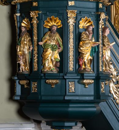 Zdjęcie nr 4: Ambona przyścienna, wisząca, z dostępem w formie jednobiegowych schodów. Kosz na planie ściętego ośmioboku, z podwieszeniem w formie spłaszczonego, ośmiobocznego wałka, zakończonego szyszką. Kosz na wysokim cokole, ścianki wydzielone pilastrami dekorowanymi muszlami i zwisami kwiatowymi, dźwigającymi pełne belkowanie z silnie wysuniętym gzymsem tworzącym parapet, dekorowanym ornamentem roślinnym. W polu ścianek nisze z konchami i figurami czterech ewangelistów, stojącymi na wspornikach. Balustrada schodów pełna, dekorowana płycinami wypełnionymi akantem, zakończona filarem z wazonem. Zaplecek zakończony gzymsem, ujęty uszami utworzonymi z suchego akantu. W polu płycina w formie stojącego prostokąta o wklęsłych narożach, w niej nisza z konchą, na tle której na wsporniku ustawiona figura Chrystusa. Baldachim na planie ściętego ośmioboku, w formie belkowania, wyłamanego na narożach. Na nim, na niskiej podstawie osiem kabłąków dekorowanych cokolikami z wazonami, podtrzymujących spłaszczoną kulę, na której stoi figura anioła z banderolą z napisem „KTO WAS SŁUCHA MNIE SŁUCHA”. Na podniebieniu baldachimu rzeźba gołębicy Ducha Świętego w glorii promienistej. Struktura polichromowana w kolorze zielonym, ze złoconymi profilami i ornamentyką. 

Figura anioła ukazana frontalnie, w kontrapoście, z głową skierowaną w lewo, obiema rękami trzyma banderolę. Twarz ma pociągłą, o migdałowatych oczach, dużym nosie i rozchylonych ustach, okoloną krótkimi, kręconymi, rozwianymi włosami. Anioł ubrany jest w długą, silnie drapowaną, złoconą tunikę, przepasaną w talii, odsłaniającą prawe ramię oraz obie nogi. Skrzydła ma złożone po bokach postaci. 

Figura Chrystusa ukazana frontalnie, w kontrapoście, prawą rękę unosi w geście błogosławieństwa, w lewej trzyma jabłko królewskie. Twarz ma szczupłą, o migdałowatych oczach, dużym nosie i wąskich ustach, okoloną brązową brodą i włosami sięgającymi ramion. Chrystus ubrany jest w przylegającą do ciała, złoconą tunikę, przepasaną w talii oraz złocony płaszcz o czerwonej podszewce, przerzucony diagonalnie przez lewe ramię. 

Figura św. Jana ukazana frontalnie, w kontrapoście, przechylona w lewo, prawą rękę wyciąga do przodu, lewą podtrzymuje otwartą księgę. Twarz ma owalną, o migdałowatych oczach, dużym nosie i wąskich ustach, okoloną brązowymi włosami sięgającymi ramion. Święty ubrany jest w przylegającą do ciała, złoconą tunikę, przepasaną w talii oraz złocony płaszcz o czerwonej podszewce, przerzucony diagonalnie przez lewe ramię, zawieszony na taśmie z przodu postaci. 

Figura św. Łukasza ukazana frontalnie, w kontrapoście, przechylona w lewo, prawą rękę unosi na tle piersi, lewą podtrzymuje otwartą księgę. Twarz ma owalną, o migdałowatych oczach, dużym nosie i wąskich ustach, okoloną brązową długą brodą i krótkimi włosami. Święty ubrany jest w przylegającą do ciała, złoconą tunikę, przepasaną w talii oraz złocony płaszcz o zielonej podszewce, przerzucony diagonalnie przez lewe ramię. Przy prawej nodze świętego głowa wołu. 

Figura św. Marka ukazana frontalnie, w kontrapoście, przechylona w lewo, z głową zwróconą w lewo, prawą rękę wyciąga w bok, lewą przytrzymuje przy boku księgę. Twarz ma owalną, o migdałowatych oczach, dużym nosie i wąskich ustach, okoloną gęstą, brązową brodą i krótkimi, kręconymi włosami. Święty ubrany jest w przylegającą do ciała, złoconą tunikę, przepasaną w talii oraz złocony płaszcz o jasnozielonej podszewce, zarzucony na ramiona i zawiązany z przodu. Przy lewej nodze postaci głowa lwa. 

Figura św. Mateusza ukazana frontalnie, w kontrapoście, lewą rękę wyciąga w bok, prawą przytrzymuje przy boku księgę. Twarz ma owalną, o migdałowatych oczach, dużym nosie i wąskich ustach, okoloną długą, brązową brodą i krótkimi włosami. Święty ubrany jest w przylegającą do ciała, złoconą tunikę, przepasaną w talii szarfą oraz złocony płaszcz o zielonej podszewce, zarzucony na ramiona i opadający po bokach. Przed świętym figurka aniołka trzymającego miskę. 
