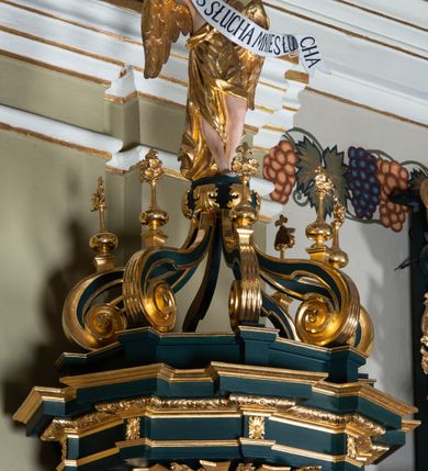 Zdjęcie nr 8: Ambona przyścienna, wisząca, z dostępem w formie jednobiegowych schodów. Kosz na planie ściętego ośmioboku, z podwieszeniem w formie spłaszczonego, ośmiobocznego wałka, zakończonego szyszką. Kosz na wysokim cokole, ścianki wydzielone pilastrami dekorowanymi muszlami i zwisami kwiatowymi, dźwigającymi pełne belkowanie z silnie wysuniętym gzymsem tworzącym parapet, dekorowanym ornamentem roślinnym. W polu ścianek nisze z konchami i figurami czterech ewangelistów, stojącymi na wspornikach. Balustrada schodów pełna, dekorowana płycinami wypełnionymi akantem, zakończona filarem z wazonem. Zaplecek zakończony gzymsem, ujęty uszami utworzonymi z suchego akantu. W polu płycina w formie stojącego prostokąta o wklęsłych narożach, w niej nisza z konchą, na tle której na wsporniku ustawiona figura Chrystusa. Baldachim na planie ściętego ośmioboku, w formie belkowania, wyłamanego na narożach. Na nim, na niskiej podstawie osiem kabłąków dekorowanych cokolikami z wazonami, podtrzymujących spłaszczoną kulę, na której stoi figura anioła z banderolą z napisem „KTO WAS SŁUCHA MNIE SŁUCHA”. Na podniebieniu baldachimu rzeźba gołębicy Ducha Świętego w glorii promienistej. Struktura polichromowana w kolorze zielonym, ze złoconymi profilami i ornamentyką. 

Figura anioła ukazana frontalnie, w kontrapoście, z głową skierowaną w lewo, obiema rękami trzyma banderolę. Twarz ma pociągłą, o migdałowatych oczach, dużym nosie i rozchylonych ustach, okoloną krótkimi, kręconymi, rozwianymi włosami. Anioł ubrany jest w długą, silnie drapowaną, złoconą tunikę, przepasaną w talii, odsłaniającą prawe ramię oraz obie nogi. Skrzydła ma złożone po bokach postaci. 

Figura Chrystusa ukazana frontalnie, w kontrapoście, prawą rękę unosi w geście błogosławieństwa, w lewej trzyma jabłko królewskie. Twarz ma szczupłą, o migdałowatych oczach, dużym nosie i wąskich ustach, okoloną brązową brodą i włosami sięgającymi ramion. Chrystus ubrany jest w przylegającą do ciała, złoconą tunikę, przepasaną w talii oraz złocony płaszcz o czerwonej podszewce, przerzucony diagonalnie przez lewe ramię. 

Figura św. Jana ukazana frontalnie, w kontrapoście, przechylona w lewo, prawą rękę wyciąga do przodu, lewą podtrzymuje otwartą księgę. Twarz ma owalną, o migdałowatych oczach, dużym nosie i wąskich ustach, okoloną brązowymi włosami sięgającymi ramion. Święty ubrany jest w przylegającą do ciała, złoconą tunikę, przepasaną w talii oraz złocony płaszcz o czerwonej podszewce, przerzucony diagonalnie przez lewe ramię, zawieszony na taśmie z przodu postaci. 

Figura św. Łukasza ukazana frontalnie, w kontrapoście, przechylona w lewo, prawą rękę unosi na tle piersi, lewą podtrzymuje otwartą księgę. Twarz ma owalną, o migdałowatych oczach, dużym nosie i wąskich ustach, okoloną brązową długą brodą i krótkimi włosami. Święty ubrany jest w przylegającą do ciała, złoconą tunikę, przepasaną w talii oraz złocony płaszcz o zielonej podszewce, przerzucony diagonalnie przez lewe ramię. Przy prawej nodze świętego głowa wołu. 

Figura św. Marka ukazana frontalnie, w kontrapoście, przechylona w lewo, z głową zwróconą w lewo, prawą rękę wyciąga w bok, lewą przytrzymuje przy boku księgę. Twarz ma owalną, o migdałowatych oczach, dużym nosie i wąskich ustach, okoloną gęstą, brązową brodą i krótkimi, kręconymi włosami. Święty ubrany jest w przylegającą do ciała, złoconą tunikę, przepasaną w talii oraz złocony płaszcz o jasnozielonej podszewce, zarzucony na ramiona i zawiązany z przodu. Przy lewej nodze postaci głowa lwa. 

Figura św. Mateusza ukazana frontalnie, w kontrapoście, lewą rękę wyciąga w bok, prawą przytrzymuje przy boku księgę. Twarz ma owalną, o migdałowatych oczach, dużym nosie i wąskich ustach, okoloną długą, brązową brodą i krótkimi włosami. Święty ubrany jest w przylegającą do ciała, złoconą tunikę, przepasaną w talii szarfą oraz złocony płaszcz o zielonej podszewce, zarzucony na ramiona i opadający po bokach. Przed świętym figurka aniołka trzymającego miskę. 
