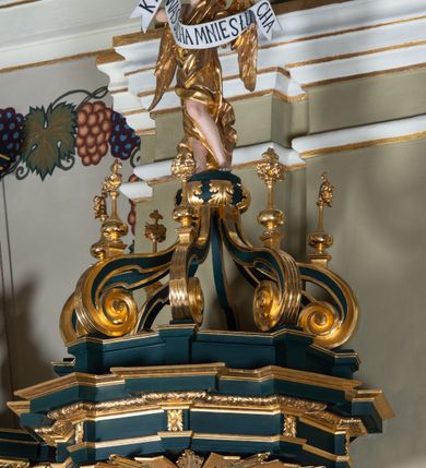 Zdjęcie nr 7: Ambona przyścienna, wisząca, z dostępem w formie jednobiegowych schodów. Kosz na planie ściętego ośmioboku, z podwieszeniem w formie spłaszczonego, ośmiobocznego wałka, zakończonego szyszką. Kosz na wysokim cokole, ścianki wydzielone pilastrami dekorowanymi muszlami i zwisami kwiatowymi, dźwigającymi pełne belkowanie z silnie wysuniętym gzymsem tworzącym parapet, dekorowanym ornamentem roślinnym. W polu ścianek nisze z konchami i figurami czterech ewangelistów, stojącymi na wspornikach. Balustrada schodów pełna, dekorowana płycinami wypełnionymi akantem, zakończona filarem z wazonem. Zaplecek zakończony gzymsem, ujęty uszami utworzonymi z suchego akantu. W polu płycina w formie stojącego prostokąta o wklęsłych narożach, w niej nisza z konchą, na tle której na wsporniku ustawiona figura Chrystusa. Baldachim na planie ściętego ośmioboku, w formie belkowania, wyłamanego na narożach. Na nim, na niskiej podstawie osiem kabłąków dekorowanych cokolikami z wazonami, podtrzymujących spłaszczoną kulę, na której stoi figura anioła z banderolą z napisem „KTO WAS SŁUCHA MNIE SŁUCHA”. Na podniebieniu baldachimu rzeźba gołębicy Ducha Świętego w glorii promienistej. Struktura polichromowana w kolorze zielonym, ze złoconymi profilami i ornamentyką. 

Figura anioła ukazana frontalnie, w kontrapoście, z głową skierowaną w lewo, obiema rękami trzyma banderolę. Twarz ma pociągłą, o migdałowatych oczach, dużym nosie i rozchylonych ustach, okoloną krótkimi, kręconymi, rozwianymi włosami. Anioł ubrany jest w długą, silnie drapowaną, złoconą tunikę, przepasaną w talii, odsłaniającą prawe ramię oraz obie nogi. Skrzydła ma złożone po bokach postaci. 

Figura Chrystusa ukazana frontalnie, w kontrapoście, prawą rękę unosi w geście błogosławieństwa, w lewej trzyma jabłko królewskie. Twarz ma szczupłą, o migdałowatych oczach, dużym nosie i wąskich ustach, okoloną brązową brodą i włosami sięgającymi ramion. Chrystus ubrany jest w przylegającą do ciała, złoconą tunikę, przepasaną w talii oraz złocony płaszcz o czerwonej podszewce, przerzucony diagonalnie przez lewe ramię. 

Figura św. Jana ukazana frontalnie, w kontrapoście, przechylona w lewo, prawą rękę wyciąga do przodu, lewą podtrzymuje otwartą księgę. Twarz ma owalną, o migdałowatych oczach, dużym nosie i wąskich ustach, okoloną brązowymi włosami sięgającymi ramion. Święty ubrany jest w przylegającą do ciała, złoconą tunikę, przepasaną w talii oraz złocony płaszcz o czerwonej podszewce, przerzucony diagonalnie przez lewe ramię, zawieszony na taśmie z przodu postaci. 

Figura św. Łukasza ukazana frontalnie, w kontrapoście, przechylona w lewo, prawą rękę unosi na tle piersi, lewą podtrzymuje otwartą księgę. Twarz ma owalną, o migdałowatych oczach, dużym nosie i wąskich ustach, okoloną brązową długą brodą i krótkimi włosami. Święty ubrany jest w przylegającą do ciała, złoconą tunikę, przepasaną w talii oraz złocony płaszcz o zielonej podszewce, przerzucony diagonalnie przez lewe ramię. Przy prawej nodze świętego głowa wołu. 

Figura św. Marka ukazana frontalnie, w kontrapoście, przechylona w lewo, z głową zwróconą w lewo, prawą rękę wyciąga w bok, lewą przytrzymuje przy boku księgę. Twarz ma owalną, o migdałowatych oczach, dużym nosie i wąskich ustach, okoloną gęstą, brązową brodą i krótkimi, kręconymi włosami. Święty ubrany jest w przylegającą do ciała, złoconą tunikę, przepasaną w talii oraz złocony płaszcz o jasnozielonej podszewce, zarzucony na ramiona i zawiązany z przodu. Przy lewej nodze postaci głowa lwa. 

Figura św. Mateusza ukazana frontalnie, w kontrapoście, lewą rękę wyciąga w bok, prawą przytrzymuje przy boku księgę. Twarz ma owalną, o migdałowatych oczach, dużym nosie i wąskich ustach, okoloną długą, brązową brodą i krótkimi włosami. Święty ubrany jest w przylegającą do ciała, złoconą tunikę, przepasaną w talii szarfą oraz złocony płaszcz o zielonej podszewce, zarzucony na ramiona i opadający po bokach. Przed świętym figurka aniołka trzymającego miskę. 
