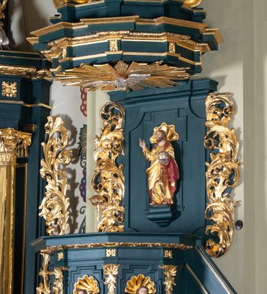 Zdjęcie nr 6: Ambona przyścienna, wisząca, z dostępem w formie jednobiegowych schodów. Kosz na planie ściętego ośmioboku, z podwieszeniem w formie spłaszczonego, ośmiobocznego wałka, zakończonego szyszką. Kosz na wysokim cokole, ścianki wydzielone pilastrami dekorowanymi muszlami i zwisami kwiatowymi, dźwigającymi pełne belkowanie z silnie wysuniętym gzymsem tworzącym parapet, dekorowanym ornamentem roślinnym. W polu ścianek nisze z konchami i figurami czterech ewangelistów, stojącymi na wspornikach. Balustrada schodów pełna, dekorowana płycinami wypełnionymi akantem, zakończona filarem z wazonem. Zaplecek zakończony gzymsem, ujęty uszami utworzonymi z suchego akantu. W polu płycina w formie stojącego prostokąta o wklęsłych narożach, w niej nisza z konchą, na tle której na wsporniku ustawiona figura Chrystusa. Baldachim na planie ściętego ośmioboku, w formie belkowania, wyłamanego na narożach. Na nim, na niskiej podstawie osiem kabłąków dekorowanych cokolikami z wazonami, podtrzymujących spłaszczoną kulę, na której stoi figura anioła z banderolą z napisem „KTO WAS SŁUCHA MNIE SŁUCHA”. Na podniebieniu baldachimu rzeźba gołębicy Ducha Świętego w glorii promienistej. Struktura polichromowana w kolorze zielonym, ze złoconymi profilami i ornamentyką. 

Figura anioła ukazana frontalnie, w kontrapoście, z głową skierowaną w lewo, obiema rękami trzyma banderolę. Twarz ma pociągłą, o migdałowatych oczach, dużym nosie i rozchylonych ustach, okoloną krótkimi, kręconymi, rozwianymi włosami. Anioł ubrany jest w długą, silnie drapowaną, złoconą tunikę, przepasaną w talii, odsłaniającą prawe ramię oraz obie nogi. Skrzydła ma złożone po bokach postaci. 

Figura Chrystusa ukazana frontalnie, w kontrapoście, prawą rękę unosi w geście błogosławieństwa, w lewej trzyma jabłko królewskie. Twarz ma szczupłą, o migdałowatych oczach, dużym nosie i wąskich ustach, okoloną brązową brodą i włosami sięgającymi ramion. Chrystus ubrany jest w przylegającą do ciała, złoconą tunikę, przepasaną w talii oraz złocony płaszcz o czerwonej podszewce, przerzucony diagonalnie przez lewe ramię. 

Figura św. Jana ukazana frontalnie, w kontrapoście, przechylona w lewo, prawą rękę wyciąga do przodu, lewą podtrzymuje otwartą księgę. Twarz ma owalną, o migdałowatych oczach, dużym nosie i wąskich ustach, okoloną brązowymi włosami sięgającymi ramion. Święty ubrany jest w przylegającą do ciała, złoconą tunikę, przepasaną w talii oraz złocony płaszcz o czerwonej podszewce, przerzucony diagonalnie przez lewe ramię, zawieszony na taśmie z przodu postaci. 

Figura św. Łukasza ukazana frontalnie, w kontrapoście, przechylona w lewo, prawą rękę unosi na tle piersi, lewą podtrzymuje otwartą księgę. Twarz ma owalną, o migdałowatych oczach, dużym nosie i wąskich ustach, okoloną brązową długą brodą i krótkimi włosami. Święty ubrany jest w przylegającą do ciała, złoconą tunikę, przepasaną w talii oraz złocony płaszcz o zielonej podszewce, przerzucony diagonalnie przez lewe ramię. Przy prawej nodze świętego głowa wołu. 

Figura św. Marka ukazana frontalnie, w kontrapoście, przechylona w lewo, z głową zwróconą w lewo, prawą rękę wyciąga w bok, lewą przytrzymuje przy boku księgę. Twarz ma owalną, o migdałowatych oczach, dużym nosie i wąskich ustach, okoloną gęstą, brązową brodą i krótkimi, kręconymi włosami. Święty ubrany jest w przylegającą do ciała, złoconą tunikę, przepasaną w talii oraz złocony płaszcz o jasnozielonej podszewce, zarzucony na ramiona i zawiązany z przodu. Przy lewej nodze postaci głowa lwa. 

Figura św. Mateusza ukazana frontalnie, w kontrapoście, lewą rękę wyciąga w bok, prawą przytrzymuje przy boku księgę. Twarz ma owalną, o migdałowatych oczach, dużym nosie i wąskich ustach, okoloną długą, brązową brodą i krótkimi włosami. Święty ubrany jest w przylegającą do ciała, złoconą tunikę, przepasaną w talii szarfą oraz złocony płaszcz o zielonej podszewce, zarzucony na ramiona i opadający po bokach. Przed świętym figurka aniołka trzymającego miskę. 
