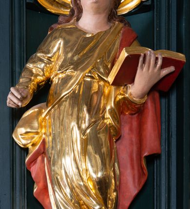 Zdjęcie nr 12: Ambona przyścienna, wisząca, z dostępem w formie jednobiegowych schodów. Kosz na planie ściętego ośmioboku, z podwieszeniem w formie spłaszczonego, ośmiobocznego wałka, zakończonego szyszką. Kosz na wysokim cokole, ścianki wydzielone pilastrami dekorowanymi muszlami i zwisami kwiatowymi, dźwigającymi pełne belkowanie z silnie wysuniętym gzymsem tworzącym parapet, dekorowanym ornamentem roślinnym. W polu ścianek nisze z konchami i figurami czterech ewangelistów, stojącymi na wspornikach. Balustrada schodów pełna, dekorowana płycinami wypełnionymi akantem, zakończona filarem z wazonem. Zaplecek zakończony gzymsem, ujęty uszami utworzonymi z suchego akantu. W polu płycina w formie stojącego prostokąta o wklęsłych narożach, w niej nisza z konchą, na tle której na wsporniku ustawiona figura Chrystusa. Baldachim na planie ściętego ośmioboku, w formie belkowania, wyłamanego na narożach. Na nim, na niskiej podstawie osiem kabłąków dekorowanych cokolikami z wazonami, podtrzymujących spłaszczoną kulę, na której stoi figura anioła z banderolą z napisem „KTO WAS SŁUCHA MNIE SŁUCHA”. Na podniebieniu baldachimu rzeźba gołębicy Ducha Świętego w glorii promienistej. Struktura polichromowana w kolorze zielonym, ze złoconymi profilami i ornamentyką. 

Figura anioła ukazana frontalnie, w kontrapoście, z głową skierowaną w lewo, obiema rękami trzyma banderolę. Twarz ma pociągłą, o migdałowatych oczach, dużym nosie i rozchylonych ustach, okoloną krótkimi, kręconymi, rozwianymi włosami. Anioł ubrany jest w długą, silnie drapowaną, złoconą tunikę, przepasaną w talii, odsłaniającą prawe ramię oraz obie nogi. Skrzydła ma złożone po bokach postaci. 

Figura Chrystusa ukazana frontalnie, w kontrapoście, prawą rękę unosi w geście błogosławieństwa, w lewej trzyma jabłko królewskie. Twarz ma szczupłą, o migdałowatych oczach, dużym nosie i wąskich ustach, okoloną brązową brodą i włosami sięgającymi ramion. Chrystus ubrany jest w przylegającą do ciała, złoconą tunikę, przepasaną w talii oraz złocony płaszcz o czerwonej podszewce, przerzucony diagonalnie przez lewe ramię. 

Figura św. Jana ukazana frontalnie, w kontrapoście, przechylona w lewo, prawą rękę wyciąga do przodu, lewą podtrzymuje otwartą księgę. Twarz ma owalną, o migdałowatych oczach, dużym nosie i wąskich ustach, okoloną brązowymi włosami sięgającymi ramion. Święty ubrany jest w przylegającą do ciała, złoconą tunikę, przepasaną w talii oraz złocony płaszcz o czerwonej podszewce, przerzucony diagonalnie przez lewe ramię, zawieszony na taśmie z przodu postaci. 

Figura św. Łukasza ukazana frontalnie, w kontrapoście, przechylona w lewo, prawą rękę unosi na tle piersi, lewą podtrzymuje otwartą księgę. Twarz ma owalną, o migdałowatych oczach, dużym nosie i wąskich ustach, okoloną brązową długą brodą i krótkimi włosami. Święty ubrany jest w przylegającą do ciała, złoconą tunikę, przepasaną w talii oraz złocony płaszcz o zielonej podszewce, przerzucony diagonalnie przez lewe ramię. Przy prawej nodze świętego głowa wołu. 

Figura św. Marka ukazana frontalnie, w kontrapoście, przechylona w lewo, z głową zwróconą w lewo, prawą rękę wyciąga w bok, lewą przytrzymuje przy boku księgę. Twarz ma owalną, o migdałowatych oczach, dużym nosie i wąskich ustach, okoloną gęstą, brązową brodą i krótkimi, kręconymi włosami. Święty ubrany jest w przylegającą do ciała, złoconą tunikę, przepasaną w talii oraz złocony płaszcz o jasnozielonej podszewce, zarzucony na ramiona i zawiązany z przodu. Przy lewej nodze postaci głowa lwa. 

Figura św. Mateusza ukazana frontalnie, w kontrapoście, lewą rękę wyciąga w bok, prawą przytrzymuje przy boku księgę. Twarz ma owalną, o migdałowatych oczach, dużym nosie i wąskich ustach, okoloną długą, brązową brodą i krótkimi włosami. Święty ubrany jest w przylegającą do ciała, złoconą tunikę, przepasaną w talii szarfą oraz złocony płaszcz o zielonej podszewce, zarzucony na ramiona i opadający po bokach. Przed świętym figurka aniołka trzymającego miskę. 
