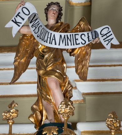 Zdjęcie nr 9: Ambona przyścienna, wisząca, z dostępem w formie jednobiegowych schodów. Kosz na planie ściętego ośmioboku, z podwieszeniem w formie spłaszczonego, ośmiobocznego wałka, zakończonego szyszką. Kosz na wysokim cokole, ścianki wydzielone pilastrami dekorowanymi muszlami i zwisami kwiatowymi, dźwigającymi pełne belkowanie z silnie wysuniętym gzymsem tworzącym parapet, dekorowanym ornamentem roślinnym. W polu ścianek nisze z konchami i figurami czterech ewangelistów, stojącymi na wspornikach. Balustrada schodów pełna, dekorowana płycinami wypełnionymi akantem, zakończona filarem z wazonem. Zaplecek zakończony gzymsem, ujęty uszami utworzonymi z suchego akantu. W polu płycina w formie stojącego prostokąta o wklęsłych narożach, w niej nisza z konchą, na tle której na wsporniku ustawiona figura Chrystusa. Baldachim na planie ściętego ośmioboku, w formie belkowania, wyłamanego na narożach. Na nim, na niskiej podstawie osiem kabłąków dekorowanych cokolikami z wazonami, podtrzymujących spłaszczoną kulę, na której stoi figura anioła z banderolą z napisem „KTO WAS SŁUCHA MNIE SŁUCHA”. Na podniebieniu baldachimu rzeźba gołębicy Ducha Świętego w glorii promienistej. Struktura polichromowana w kolorze zielonym, ze złoconymi profilami i ornamentyką. 

Figura anioła ukazana frontalnie, w kontrapoście, z głową skierowaną w lewo, obiema rękami trzyma banderolę. Twarz ma pociągłą, o migdałowatych oczach, dużym nosie i rozchylonych ustach, okoloną krótkimi, kręconymi, rozwianymi włosami. Anioł ubrany jest w długą, silnie drapowaną, złoconą tunikę, przepasaną w talii, odsłaniającą prawe ramię oraz obie nogi. Skrzydła ma złożone po bokach postaci. 

Figura Chrystusa ukazana frontalnie, w kontrapoście, prawą rękę unosi w geście błogosławieństwa, w lewej trzyma jabłko królewskie. Twarz ma szczupłą, o migdałowatych oczach, dużym nosie i wąskich ustach, okoloną brązową brodą i włosami sięgającymi ramion. Chrystus ubrany jest w przylegającą do ciała, złoconą tunikę, przepasaną w talii oraz złocony płaszcz o czerwonej podszewce, przerzucony diagonalnie przez lewe ramię. 

Figura św. Jana ukazana frontalnie, w kontrapoście, przechylona w lewo, prawą rękę wyciąga do przodu, lewą podtrzymuje otwartą księgę. Twarz ma owalną, o migdałowatych oczach, dużym nosie i wąskich ustach, okoloną brązowymi włosami sięgającymi ramion. Święty ubrany jest w przylegającą do ciała, złoconą tunikę, przepasaną w talii oraz złocony płaszcz o czerwonej podszewce, przerzucony diagonalnie przez lewe ramię, zawieszony na taśmie z przodu postaci. 

Figura św. Łukasza ukazana frontalnie, w kontrapoście, przechylona w lewo, prawą rękę unosi na tle piersi, lewą podtrzymuje otwartą księgę. Twarz ma owalną, o migdałowatych oczach, dużym nosie i wąskich ustach, okoloną brązową długą brodą i krótkimi włosami. Święty ubrany jest w przylegającą do ciała, złoconą tunikę, przepasaną w talii oraz złocony płaszcz o zielonej podszewce, przerzucony diagonalnie przez lewe ramię. Przy prawej nodze świętego głowa wołu. 

Figura św. Marka ukazana frontalnie, w kontrapoście, przechylona w lewo, z głową zwróconą w lewo, prawą rękę wyciąga w bok, lewą przytrzymuje przy boku księgę. Twarz ma owalną, o migdałowatych oczach, dużym nosie i wąskich ustach, okoloną gęstą, brązową brodą i krótkimi, kręconymi włosami. Święty ubrany jest w przylegającą do ciała, złoconą tunikę, przepasaną w talii oraz złocony płaszcz o jasnozielonej podszewce, zarzucony na ramiona i zawiązany z przodu. Przy lewej nodze postaci głowa lwa. 

Figura św. Mateusza ukazana frontalnie, w kontrapoście, lewą rękę wyciąga w bok, prawą przytrzymuje przy boku księgę. Twarz ma owalną, o migdałowatych oczach, dużym nosie i wąskich ustach, okoloną długą, brązową brodą i krótkimi włosami. Święty ubrany jest w przylegającą do ciała, złoconą tunikę, przepasaną w talii szarfą oraz złocony płaszcz o zielonej podszewce, zarzucony na ramiona i opadający po bokach. Przed świętym figurka aniołka trzymającego miskę. 
