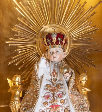 Zdjęcie nr 2: Figurka nagiego Dzieciątka przeznaczonego do ubierania. Jezus ukazany w pozycji siedzącej, frontalnie, z głową delikatnie skierowaną w prawo, lewą rękę wyciąga przed siebie, prawą, ugiętą, unosi w geście błogosławieństwa. Twarz ma kwadratową, o pełnych policzkach, wysokim czole i dużych uszach. Oczy ma migdałowate, nos mały, zadarty, usta niewielkie, z podkreśloną brodą. Twarz okolona krótkimi, kręconymi włosami. Ciało szczupłe, o delikatnie zarysowanej klatce piersiowej. Dzieciątko ubrane jest w bogate szaty z tkaniny o haftowanym wzorze, dekorowane koralikami: białą tunikę i złoty płaszcz, w lewej dłoni trzyma jabłko królewskie, na głowie ma nałożoną koronę zamkniętą, a na szyi zawieszone perły. Figurka posadzona jest na złoconym tronie o podłokietnikach w formie herm oraz zaplecku zwieńczonym złoconym medalionem w glorii promienistej. Tron ustawiony jest na złoconym cokole zakończonym gzymsem, z rytym monogramem IHS, flankowanym motywem winnego grona. 