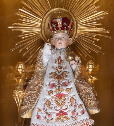 Zdjęcie nr 1: Figurka nagiego Dzieciątka przeznaczonego do ubierania. Jezus ukazany w pozycji siedzącej, frontalnie, z głową delikatnie skierowaną w prawo, lewą rękę wyciąga przed siebie, prawą, ugiętą, unosi w geście błogosławieństwa. Twarz ma kwadratową, o pełnych policzkach, wysokim czole i dużych uszach. Oczy ma migdałowate, nos mały, zadarty, usta niewielkie, z podkreśloną brodą. Twarz okolona krótkimi, kręconymi włosami. Ciało szczupłe, o delikatnie zarysowanej klatce piersiowej. Dzieciątko ubrane jest w bogate szaty z tkaniny o haftowanym wzorze, dekorowane koralikami: białą tunikę i złoty płaszcz, w lewej dłoni trzyma jabłko królewskie, na głowie ma nałożoną koronę zamkniętą, a na szyi zawieszone perły. Figurka posadzona jest na złoconym tronie o podłokietnikach w formie herm oraz zaplecku zwieńczonym złoconym medalionem w glorii promienistej. Tron ustawiony jest na złoconym cokole zakończonym gzymsem, z rytym monogramem IHS, flankowanym motywem winnego grona. 