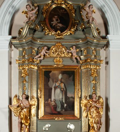 Zdjęcie nr 30: Ołtarz jednoosiowy, jednokondygnacyjny ze zwieńczeniem. Podwójny cokół z wąską, drewnianą mensą ołtarzową, tabernakulum ze spływami wolutowymi. Retabulum flankowane dwiema ćwierć kolumnami skierowanymi po ukosie na zewnątrz, z których każda ujęta parą korynckich, kanelowanych pilastrów dźwigających przełamujące się nad nimi belkowanie i dwa faliste naczółki. Na tle pilastrów, na konsolach stoją dwa uskrzydlone anioły. Zwieńczenie ujęte spływami wolutowymi, zakończone dwiema profilowanym, lekko falistym gzymsem z dwiema stykającymi się wolutami. W polu głównym szeroka, złocona, prostokątna rama z obrazami „Św. Mikołaj” i "Wizja św. Franciszka" na zasuwie, w zwieńczeniu owalny obraz w złoconej ramie z przedstawieniem św. Antoniego. Figury aniołów wydrążone, ukazane w kontrapoście, z dłońmi wysuniętymi do przodu; anioł po lewej trzyma chustę. Ich twarze są owalne o regularnych rysach, z brązowymi puklowanymi włosami sięgającymi ramion. Z tyłu przymocowane duże, złote skrzydła. Ubrane są w długie, złote szaty o ostrym zarysie, odsłaniające klatkę piersiową i jedną z nóg. W partii belkowania para nagich puttów ze złoconymi opaskami na biodrach ujmujących kartusz w rokokowym obramieniu z chrystogramem. Na naczółkach z kolei siedzą nagie putta z przepaskami na biodrach i złotymi skrzydełkami. Na dekorację ornamentalną składają się drobne motywy roślinne, kampanule na ćwierćkolumnach, zwisy róż i gałązek wokół obrazu w zwieńczeniu i efektowna podwójna muszla palmetowa w zwieńczeniu. 
