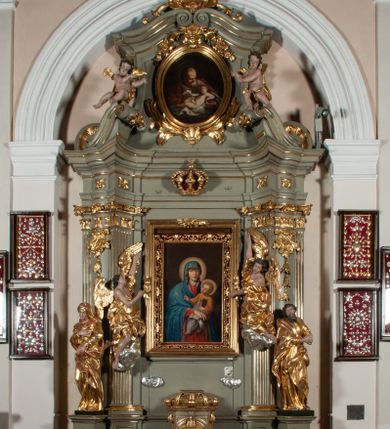 Zdjęcie nr 1: Ołtarz jednoosiowy, jednokondygnacyjny ze zwieńczeniem. Podwójny cokół z wąską, drewnianą mensą ołtarzową, wybrzuszoną. Na niej stoi tabernakulum ujęte narożnie ustawionymi spływami wolutowymi, zwieńczone spłaszczoną kopułką ze spływami wolutowymi. Retabulum flankowane dwiema ćwierć kolumnami skierowanymi po ukosie na zewnątrz, z których każda ujęta parą korynckich, kanelowanych pilastrów dźwigających przełamujące się nad nimi belkowanie i dwa faliste naczółki. Na tle pilastrów, na konsolach stoją figury św. Anny i św. Joachima oraz unoszą się na chmurach flankujące obraz uskrzydlone anioły. Zwieńczenie ujęte spływami wolutowymi, zakończone profilowanym, lekko falistym gzymsem z dwiema stykającymi się wolutami. N naczółkach dwa uskrzydlone, przepasane opaską na biodrach aniołki. W polu głównym szeroka, złocona, prostokątna rama z obrazem Matki Boskiej z Dzieciątkiem i koroną w partii belkowania, a w zwieńczeniu owalny obraz w złoconej ramie z przedstawieniem św. Józefa z Dzieciątkiem. Figury św. Anny i św. Joachima ukazane w lekkim kontrapoście, w długich, obficie fałdowanych szatach. Anna trzyma prawą rękę na piersi, w lewej przytrzymuje połę płaszcza, natomiast Joachim ręce składa w geście modlitwy. Anioły klęczą na chmurach, usytuowanych na różnych poziomach, ubrane są w miękko, ale drobno fałdowane szaty, odsłaniające ich ramiona i górną część klatki piersiowej i wskazujący na obraz. Na dekorację ornamentalną składają się drobne motywy roślinne, kampanule na ćwierćkolumnach, zwisy róż i gałązek palm wokół obrazu w zwieńczeniu i efektowna podwójna muszla palmetowa w zwieńczeniu. 