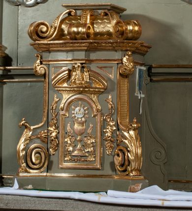 Zdjęcie nr 20: Ołtarz jednoosiowy, jednokondygnacyjny ze zwieńczeniem. Podwójny cokół z wąską, drewnianą mensą ołtarzową, wybrzuszoną. Na niej stoi tabernakulum ujęte narożnie ustawionymi spływami wolutowymi, zwieńczone spłaszczoną kopułką ze spływami wolutowymi. Retabulum flankowane dwiema ćwierć kolumnami skierowanymi po ukosie na zewnątrz, z których każda ujęta parą korynckich, kanelowanych pilastrów dźwigających przełamujące się nad nimi belkowanie i dwa faliste naczółki. Na tle pilastrów, na konsolach stoją figury św. Anny i św. Joachima oraz unoszą się na chmurach flankujące obraz uskrzydlone anioły. Zwieńczenie ujęte spływami wolutowymi, zakończone profilowanym, lekko falistym gzymsem z dwiema stykającymi się wolutami. N naczółkach dwa uskrzydlone, przepasane opaską na biodrach aniołki. W polu głównym szeroka, złocona, prostokątna rama z obrazem Matki Boskiej z Dzieciątkiem i koroną w partii belkowania, a w zwieńczeniu owalny obraz w złoconej ramie z przedstawieniem św. Józefa z Dzieciątkiem. Figury św. Anny i św. Joachima ukazane w lekkim kontrapoście, w długich, obficie fałdowanych szatach. Anna trzyma prawą rękę na piersi, w lewej przytrzymuje połę płaszcza, natomiast Joachim ręce składa w geście modlitwy. Anioły klęczą na chmurach, usytuowanych na różnych poziomach, ubrane są w miękko, ale drobno fałdowane szaty, odsłaniające ich ramiona i górną część klatki piersiowej i wskazujący na obraz. Na dekorację ornamentalną składają się drobne motywy roślinne, kampanule na ćwierćkolumnach, zwisy róż i gałązek palm wokół obrazu w zwieńczeniu i efektowna podwójna muszla palmetowa w zwieńczeniu. 