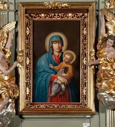 Zdjęcie nr 6: Obraz w kształcie stojącego prostokąta ukazuje na ciemnym, brązowym, jednolitym tle Matkę Boską z Dzieciątkiem na rękach. Maria ukazana jest do kolan, frontalnie, ze skrzyżowanymi rękoma, z których prawa z wyprostowanymi dwoma palcami, a lewa zaciśnięta z chustką. Jej twarz modelowana jest miękko, nos smukły, usta czerwone, uśmiechnięte, oczy otwarte, spoglądające na wprost. Ubrana jest w czerwoną suknię i niebieski płaszcz z różowym podbiciem, zarzucony na głowę. Jezus jest ukazany w trzech czwartych, zwrócony w prawo. Twarz ma pulchną, oczy szeroko otwarte, nos krótki, usta pełne, zaczerwienione, włosy jasne, kręcone, krótkie. Jest ubrany w brązową sukienkę i białą pieluchę wokół bioder i nóg. Prawą rękę unosi w kierunku gwiazdy na lewym ramieniu Marii, a w prawej trzyma księgę, na okładce której widnieją litery IHS w glorii promienistej. Wokół głowy Marii i Jezusa rozświetlone nimby. 

