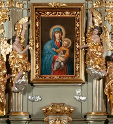 Zdjęcie nr 5: Obraz w kształcie stojącego prostokąta ukazuje na ciemnym, brązowym, jednolitym tle Matkę Boską z Dzieciątkiem na rękach. Maria ukazana jest do kolan, frontalnie, ze skrzyżowanymi rękoma, z których prawa z wyprostowanymi dwoma palcami, a lewa zaciśnięta z chustką. Jej twarz modelowana jest miękko, nos smukły, usta czerwone, uśmiechnięte, oczy otwarte, spoglądające na wprost. Ubrana jest w czerwoną suknię i niebieski płaszcz z różowym podbiciem, zarzucony na głowę. Jezus jest ukazany w trzech czwartych, zwrócony w prawo. Twarz ma pulchną, oczy szeroko otwarte, nos krótki, usta pełne, zaczerwienione, włosy jasne, kręcone, krótkie. Jest ubrany w brązową sukienkę i białą pieluchę wokół bioder i nóg. Prawą rękę unosi w kierunku gwiazdy na lewym ramieniu Marii, a w prawej trzyma księgę, na okładce której widnieją litery IHS w glorii promienistej. Wokół głowy Marii i Jezusa rozświetlone nimby. 
