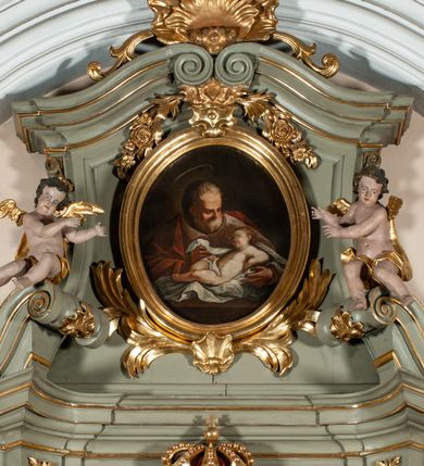 Zdjęcie nr 2: Obraz owalny ukazuje na nieokreślonym, brązowym tle św. Józefa i leżącego przed nim w pieluchach Jezusa. Święty ukazany za stołem, do pasa, ubrany w brązową suknię i czerwony płaszcz. Jego twarz zwrócona w trzech czwartych w lewo, w kierunku leżącego przed nim Jezusa, silnie oświetlona, o wysokim czole, krótkim nosie, siwej, gęstej brodzie i krótkich, siwych włosach; nad głowa widniej aureola. Leżący w niemal nagi Jezus, przepasany na biodrach pieluchą, wspierający się na ugiętej w łokciu lewej ręce, z prawą opartą na biodrze, z zgiętymi lekko nogami w kolanach. Twarz ma okragłą, pełną, nos krótki, usta drobne, lekko rozchylone, policzki zaczerwienione. Na głowie włosy krótkie, wokół niej promienie. Tło ciemne, nieokreślone.  