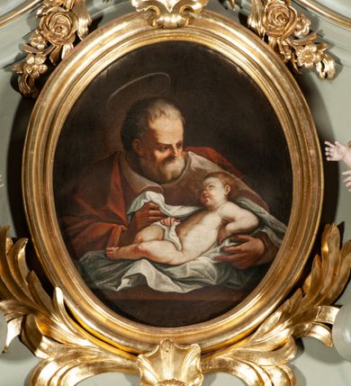 Zdjęcie nr 1: Obraz owalny ukazuje na nieokreślonym, brązowym tle św. Józefa i leżącego przed nim w pieluchach Jezusa. Święty ukazany za stołem, do pasa, ubrany w brązową suknię i czerwony płaszcz. Jego twarz zwrócona w trzech czwartych w lewo, w kierunku leżącego przed nim Jezusa, silnie oświetlona, o wysokim czole, krótkim nosie, siwej, gęstej brodzie i krótkich, siwych włosach; nad głowa widniej aureola. Leżący w niemal nagi Jezus, przepasany na biodrach pieluchą, wspierający się na ugiętej w łokciu lewej ręce, z prawą opartą na biodrze, z zgiętymi lekko nogami w kolanach. Twarz ma okragłą, pełną, nos krótki, usta drobne, lekko rozchylone, policzki zaczerwienione. Na głowie włosy krótkie, wokół niej promienie. Tło ciemne, nieokreślone.  