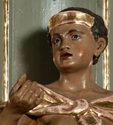 Zdjęcie nr 19: Ołtarz jednoosiowy, jednokondygnacyjny ze zwieńczeniem. Podwójny cokół z wąską, drewnianą mensą ołtarzową, wybrzuszoną. Na niej stoi tabernakulum ujęte kolumienkami i spływami wolutowymi, zwieńczone dwoma zwróconymi do siebie naczółkami z niewielką figurą baranka na tle promieni. Retabulum flankowane dwiema ćwierć kolumnami skierowanymi po ukosie na zewnątrz, z których każda ujęta parą korynckich, kanelowanych pilastrów dźwigających przełamujące się nad nimi belkowanie i dwa faliste naczółki. Na tle pilastrów, na konsolach stoją dwie figury egzotycznych mężczyzn. Zwieńczenie ujęte spływami wolutowymi, zakończone profilowanym, lekko falistym gzymsem z dwiema stykającymi się wolutami. W polu głównym szeroka, złocona, prostokątna rama z obrazem &quot;Męczeństwo św. Bartłomieja&quot;, w zwieńczeniu owalny obraz w złoconej ramie z przedstawieniem Anioła Stróża. 
Figury egzotycznych (o typach wschodnich) mężczyzn ukazane w kontrapoście, o prawidłowych proporcjach ciała, z rękami uniesionymi przed siebie, na wysokości klatki piersiowej. Ich twarze są pełne, okrągłe o szerokich nosach i wydatnych, lekko otwartych ustach, na głowie krótkie, kręcone, ciemne włosy przytrzymane na czole złoconymi opaskami. Biodra okalają sięgające kolan spódniczki ze srebrnych piór. Mężczyzna po lewej stronie trzyma skórę z twarzą św. Bartłomieja osłaniającą część jego ciała, a postać po prawej nosi na plechach płaszcz sięgający stóp. Na dekorację ornamentalną składają się drobne motywy roślinne, kampanule na ćwierćkolumnach, kartusz ujęty drobnymi rocaille’ami, zwisy róż i gałązek palm wokół obrazu w zwieńczeniu i efektowna podwójna muszla palmetowa w zwieńczeniu. 
