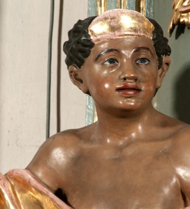 Zdjęcie nr 26: Ołtarz jednoosiowy, jednokondygnacyjny ze zwieńczeniem. Podwójny cokół z wąską, drewnianą mensą ołtarzową, wybrzuszoną. Na niej stoi tabernakulum ujęte kolumienkami i spływami wolutowymi, zwieńczone dwoma zwróconymi do siebie naczółkami z niewielką figurą baranka na tle promieni. Retabulum flankowane dwiema ćwierć kolumnami skierowanymi po ukosie na zewnątrz, z których każda ujęta parą korynckich, kanelowanych pilastrów dźwigających przełamujące się nad nimi belkowanie i dwa faliste naczółki. Na tle pilastrów, na konsolach stoją dwie figury egzotycznych mężczyzn. Zwieńczenie ujęte spływami wolutowymi, zakończone profilowanym, lekko falistym gzymsem z dwiema stykającymi się wolutami. W polu głównym szeroka, złocona, prostokątna rama z obrazem "Męczeństwo św. Bartłomieja", w zwieńczeniu owalny obraz w złoconej ramie z przedstawieniem Anioła Stróża. 
Figury egzotycznych (o typach wschodnich) mężczyzn ukazane w kontrapoście, o prawidłowych proporcjach ciała, z rękami uniesionymi przed siebie, na wysokości klatki piersiowej. Ich twarze są pełne, okrągłe o szerokich nosach i wydatnych, lekko otwartych ustach, na głowie krótkie, kręcone, ciemne włosy przytrzymane na czole złoconymi opaskami. Biodra okalają sięgające kolan spódniczki ze srebrnych piór. Mężczyzna po lewej stronie trzyma skórę z twarzą św. Bartłomieja osłaniającą część jego ciała, a postać po prawej nosi na plechach płaszcz sięgający stóp. Na dekorację ornamentalną składają się drobne motywy roślinne, kampanule na ćwierćkolumnach, kartusz ujęty drobnymi rocaille’ami, zwisy róż i gałązek palm wokół obrazu w zwieńczeniu i efektowna podwójna muszla palmetowa w zwieńczeniu. 
