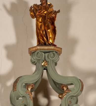 Zdjęcie nr 21: Antyambona składa się z wysokiego cokołu ujętego  szerokimi, kanelowanymi spływami wolutowymi i figurą baranka, zaplecka z obrazem ukazującym chrzest Chrystusa w Jordanie, ujętego podwieszoną kotarą i baldachim z podwieszoną pod nim kotarą. Na nim cztery esowato wygięte i łączące się górą ze sobą kabłąki, zakończone personifikacją Kościoła – kobiety w tiarze papieskiej na głowie. 