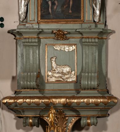 Zdjęcie nr 13: Antyambona składa się z wysokiego cokołu ujętego  szerokimi, kanelowanymi spływami wolutowymi i figurą baranka, zaplecka z obrazem ukazującym chrzest Chrystusa w Jordanie, ujętego podwieszoną kotarą i baldachim z podwieszoną pod nim kotarą. Na nim cztery esowato wygięte i łączące się górą ze sobą kabłąki, zakończone personifikacją Kościoła – kobiety w tiarze papieskiej na głowie. 