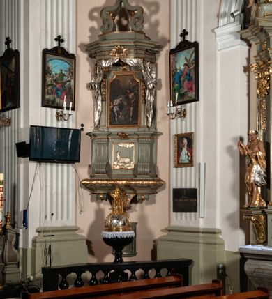 Zdjęcie nr 2: Antyambona składa się z wysokiego cokołu ujętego  szerokimi, kanelowanymi spływami wolutowymi i figurą baranka, zaplecka z obrazem ukazującym chrzest Chrystusa w Jordanie, ujętego podwieszoną kotarą i baldachim z podwieszoną pod nim kotarą. Na nim cztery esowato wygięte i łączące się górą ze sobą kabłąki, zakończone personifikacją Kościoła – kobiety w tiarze papieskiej na głowie. 
