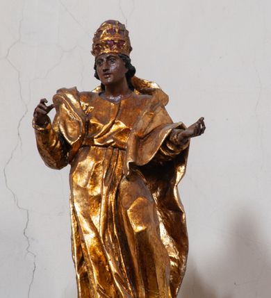 Zdjęcie nr 23: Antyambona składa się z wysokiego cokołu ujętego  szerokimi, kanelowanymi spływami wolutowymi i figurą baranka, zaplecka z obrazem ukazującym chrzest Chrystusa w Jordanie, ujętego podwieszoną kotarą i baldachim z podwieszoną pod nim kotarą. Na nim cztery esowato wygięte i łączące się górą ze sobą kabłąki, zakończone personifikacją Kościoła – kobiety w tiarze papieskiej na głowie. 