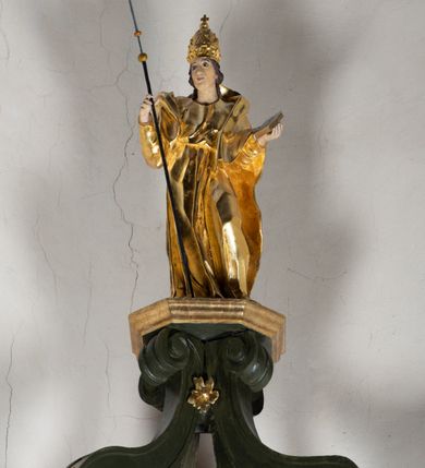 Zdjęcie nr 15: Antyambona składa się z wysokiego cokołu ujętego  szerokimi, kanelowanymi spływami wolutowymi i figurą baranka, zaplecka z obrazem ukazującym chrzest Chrystusa w Jordanie, ujętego podwieszoną kotarą i baldachim z podwieszoną pod nim kotarą. Na nim cztery esowato wygięte i łączące się górą ze sobą kabłąki, zakończone personifikacją Kościoła – kobiety w tiarze papieskiej na głowie. 