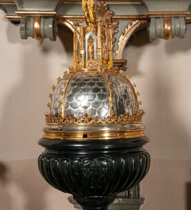 Zdjęcie nr 5: Podstawa okrągła, dwustopniowa z prostopadłościenną górną częścią, z trzonem w typie gruszkowatym z wałkiem w górnej części, podtrzymującym czarę w formie spłaszczonej kuli z puklami. Na czarze tarcza herbowa ujęta wolutami z godłem herbu Szaszor lub Orla. Pokrywa drewniana, w kształcie smukłej półkopuły o powierzchni pokrytej srebrzonymi łuskami, obwiedzionej koroną, podzielona żebrami z żabkami i zwieńczona koroną. Nad kopułą sześcioboczna wieżyczka ujęta wolnostojącymi pinaklami. Jej ścianki zakończone trójkątnie z ornamentalnymi grzebieniami, wypełnione wnękami z postaciami aniołów. Nad wieżyczką figura św. Jana Chrzciciela, ubranego w wielbłądzią skórę i płaszcz przerzucony na prawym ramieniu. W prawej dłoni podtrzymuje baranka, w lewej trzyma osadzony na długim trzpieniu krzyż. Na wewnętrznej stronie pokrywy tabliczka z inskrypcją: „CURA ET IMPENSIS / JOSEPHI PAJĄCZEWSKI / CURATI MORAVICEAEN ET / DECANI CZERNICHOVIEN A(NNO) D(OMINI) 1904 A(D) M(AIOREM) D(EI) G(LORIAM)”.        