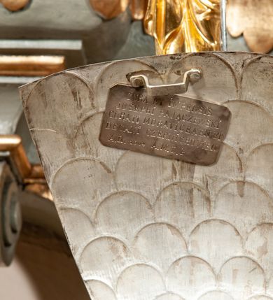 Zdjęcie nr 3: Podstawa okrągła, dwustopniowa z prostopadłościenną górną częścią, z trzonem w typie gruszkowatym z wałkiem w górnej części, podtrzymującym czarę w formie spłaszczonej kuli z puklami. Na czarze tarcza herbowa ujęta wolutami z godłem herbu Szaszor lub Orla. Pokrywa drewniana, w kształcie smukłej półkopuły o powierzchni pokrytej srebrzonymi łuskami, obwiedzionej koroną, podzielona żebrami z żabkami i zwieńczona koroną. Nad kopułą sześcioboczna wieżyczka ujęta wolnostojącymi pinaklami. Jej ścianki zakończone trójkątnie z ornamentalnymi grzebieniami, wypełnione wnękami z postaciami aniołów. Nad wieżyczką figura św. Jana Chrzciciela, ubranego w wielbłądzią skórę i płaszcz przerzucony na prawym ramieniu. W prawej dłoni podtrzymuje baranka, w lewej trzyma osadzony na długim trzpieniu krzyż. Na wewnętrznej stronie pokrywy tabliczka z inskrypcją: „CURA ET IMPENSIS / JOSEPHI PAJĄCZEWSKI / CURATI MORAVICEAEN ET / DECANI CZERNICHOVIEN A(NNO) D(OMINI) 1904 A(D) M(AIOREM) D(EI) G(LORIAM)”.        