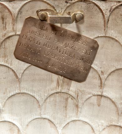 Zdjęcie nr 12: Podstawa okrągła, dwustopniowa z prostopadłościenną górną częścią, z trzonem w typie gruszkowatym z wałkiem w górnej części, podtrzymującym czarę w formie spłaszczonej kuli z puklami. Na czarze tarcza herbowa ujęta wolutami z godłem herbu Szaszor lub Orla. Pokrywa drewniana, w kształcie smukłej półkopuły o powierzchni pokrytej srebrzonymi łuskami, obwiedzionej koroną, podzielona żebrami z żabkami i zwieńczona koroną. Nad kopułą sześcioboczna wieżyczka ujęta wolnostojącymi pinaklami. Jej ścianki zakończone trójkątnie z ornamentalnymi grzebieniami, wypełnione wnękami z postaciami aniołów. Nad wieżyczką figura św. Jana Chrzciciela, ubranego w wielbłądzią skórę i płaszcz przerzucony na prawym ramieniu. W prawej dłoni podtrzymuje baranka, w lewej trzyma osadzony na długim trzpieniu krzyż. Na wewnętrznej stronie pokrywy tabliczka z inskrypcją: „CURA ET IMPENSIS / JOSEPHI PAJĄCZEWSKI / CURATI MORAVICEAEN ET / DECANI CZERNICHOVIEN A(NNO) D(OMINI) 1904 A(D) M(AIOREM) D(EI) G(LORIAM)”.        