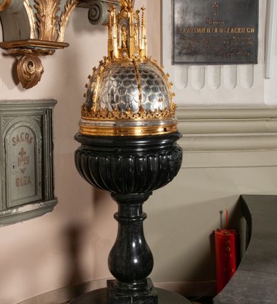 Zdjęcie nr 1: Podstawa okrągła, dwustopniowa z prostopadłościenną górną częścią, z trzonem w typie gruszkowatym z wałkiem w górnej części, podtrzymującym czarę w formie spłaszczonej kuli z puklami. Na czarze tarcza herbowa ujęta wolutami z godłem herbu Szaszor lub Orla. Pokrywa drewniana, w kształcie smukłej półkopuły o powierzchni pokrytej srebrzonymi łuskami, obwiedzionej koroną, podzielona żebrami z żabkami i zwieńczona koroną. Nad kopułą sześcioboczna wieżyczka ujęta wolnostojącymi pinaklami. Jej ścianki zakończone trójkątnie z ornamentalnymi grzebieniami, wypełnione wnękami z postaciami aniołów. Nad wieżyczką figura św. Jana Chrzciciela, ubranego w wielbłądzią skórę i płaszcz przerzucony na prawym ramieniu. W prawej dłoni podtrzymuje baranka, w lewej trzyma osadzony na długim trzpieniu krzyż. Na wewnętrznej stronie pokrywy tabliczka z inskrypcją: „CURA ET IMPENSIS / JOSEPHI PAJĄCZEWSKI / CURATI MORAVICEAEN ET / DECANI CZERNICHOVIEN A(NNO) D(OMINI) 1904 A(D) M(AIOREM) D(EI) G(LORIAM)”.        