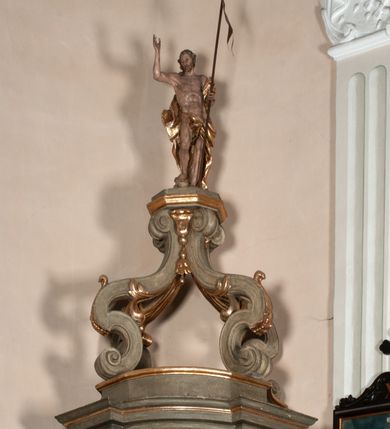 Zdjęcie nr 18: Kosz ambony na rzucie czworoboku, o bokach lekko wypukłych, wsparty na piramidalnym cokole z szyszką ujętą liśćmi. Kosz podzielony szerokimi, kanelowanymi spływami i zwieńczony gzymsem. Na ściance frontowej płaskorzeźbiona figura Chrystusa błogosławiącego trzymany w ręku bochenek chleba. Zaplecek flankowany profilowanymi listwami z kampanulami zakończonymi ulistnionymi konsolkami, pośrodku obraz w kształcie stojącego prostokąta zamkniętego łukiem półkolistym, nadwieszonym z przedstawieniem Jezusa wśród mędrców. Baldachim powielający rzut kosza, profilowany, ujęty impostami, zwieńczony czterema esowato wygiętymi i łączącymi się górą kabłąkami. Na podniebieniu baldachimu gołębica Ducha Świętego ,a tle promieni. Na nich figura Chrystusa Zmartwychwstałego, ukazanego w całej postaci, w kontrapoście, z płaszczem na plecach osłaniającym biodro. Prawą rękę unosi w geście błogosławieństwa, w lewej trzyma chorągiew. 