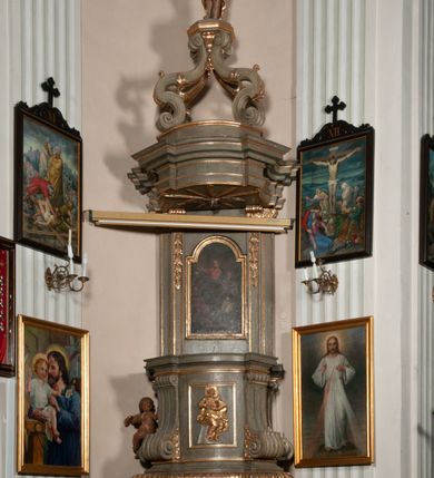 Zdjęcie nr 16: Kosz ambony na rzucie czworoboku, o bokach lekko wypukłych, wsparty na piramidalnym cokole z szyszką ujętą liśćmi. Kosz podzielony szerokimi, kanelowanymi spływami i zwieńczony gzymsem. Na ściance frontowej płaskorzeźbiona figura Chrystusa błogosławiącego trzymany w ręku bochenek chleba. Zaplecek flankowany profilowanymi listwami z kampanulami zakończonymi ulistnionymi konsolkami, pośrodku obraz w kształcie stojącego prostokąta zamkniętego łukiem półkolistym, nadwieszonym z przedstawieniem Jezusa wśród mędrców. Baldachim powielający rzut kosza, profilowany, ujęty impostami, zwieńczony czterema esowato wygiętymi i łączącymi się górą kabłąkami. Na podniebieniu baldachimu gołębica Ducha Świętego ,a tle promieni. Na nich figura Chrystusa Zmartwychwstałego, ukazanego w całej postaci, w kontrapoście, z płaszczem na plecach osłaniającym biodro. Prawą rękę unosi w geście błogosławieństwa, w lewej trzyma chorągiew. 