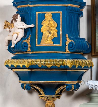 Zdjęcie nr 3: Kosz ambony na rzucie czworoboku, o bokach lekko wypukłych, wsparty na piramidalnym cokole z szyszką ujętą liśćmi. Kosz podzielony szerokimi, kanelowanymi spływami i zwieńczony gzymsem. Na ściance frontowej płaskorzeźbiona figura Chrystusa błogosławiącego trzymany w ręku bochenek chleba. Zaplecek flankowany profilowanymi listwami z kampanulami zakończonymi ulistnionymi konsolkami, pośrodku obraz w kształcie stojącego prostokąta zamkniętego łukiem półkolistym, nadwieszonym z przedstawieniem Jezusa wśród mędrców. Baldachim powielający rzut kosza, profilowany, ujęty impostami, zwieńczony czterema esowato wygiętymi i łączącymi się górą kabłąkami. Na podniebieniu baldachimu gołębica Ducha Świętego ,a tle promieni. Na nich figura Chrystusa Zmartwychwstałego, ukazanego w całej postaci, w kontrapoście, z płaszczem na plecach osłaniającym biodro. Prawą rękę unosi w geście błogosławieństwa, w lewej trzyma chorągiew. 