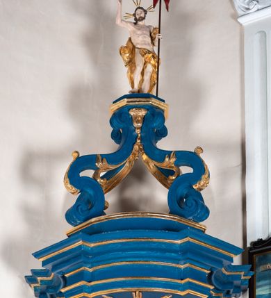 Zdjęcie nr 10: Kosz ambony na rzucie czworoboku, o bokach lekko wypukłych, wsparty na piramidalnym cokole z szyszką ujętą liśćmi. Kosz podzielony szerokimi, kanelowanymi spływami i zwieńczony gzymsem. Na ściance frontowej płaskorzeźbiona figura Chrystusa błogosławiącego trzymany w ręku bochenek chleba. Zaplecek flankowany profilowanymi listwami z kampanulami zakończonymi ulistnionymi konsolkami, pośrodku obraz w kształcie stojącego prostokąta zamkniętego łukiem półkolistym, nadwieszonym z przedstawieniem Jezusa wśród mędrców. Baldachim powielający rzut kosza, profilowany, ujęty impostami, zwieńczony czterema esowato wygiętymi i łączącymi się górą kabłąkami. Na podniebieniu baldachimu gołębica Ducha Świętego ,a tle promieni. Na nich figura Chrystusa Zmartwychwstałego, ukazanego w całej postaci, w kontrapoście, z płaszczem na plecach osłaniającym biodro. Prawą rękę unosi w geście błogosławieństwa, w lewej trzyma chorągiew. 