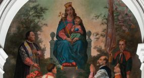 Zdjęcie nr 1: Fresk wkomponowany w arkadę zamkniętą półkoliście, w ścianie południowej prezbiterium, nad portalem do aneksu. Ukazuje siedzącą na tronie Matkę Boską z Dzieciątkiem na kolanach oraz klęczących i stojących przed nią od lewej proboszcza Józefa Krzemińskiego, rodzinę w stroju ludowym, Izabelę i Felicjana Szybalskich oraz chłopca z chorągwią w ręku. 