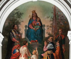 Zdjęcie nr 1: Fresk wkomponowany w arkadę zamkniętą półkoliście, w ścianie południowej prezbiterium, nad portalem do aneksu. Ukazuje siedzącą na tronie Matkę Boską z Dzieciątkiem na kolanach oraz klęczących i stojących przed nią od lewej proboszcza Józefa Krzemińskiego, rodzinę w stroju ludowym, Izabelę i Felicjana Szybalskich oraz chłopca z chorągwią w ręku. 