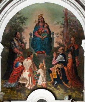 Zdjęcie nr 1: Fresk wkomponowany w arkadę zamkniętą półkoliście, w ścianie południowej prezbiterium, nad portalem do aneksu. Ukazuje siedzącą na tronie Matkę Boską z Dzieciątkiem na kolanach oraz klęczących i stojących przed nią od lewej proboszcza Józefa Krzemińskiego, rodzinę w stroju ludowym, Izabelę i Felicjana Szybalskich oraz chłopca z chorągwią w ręku. 